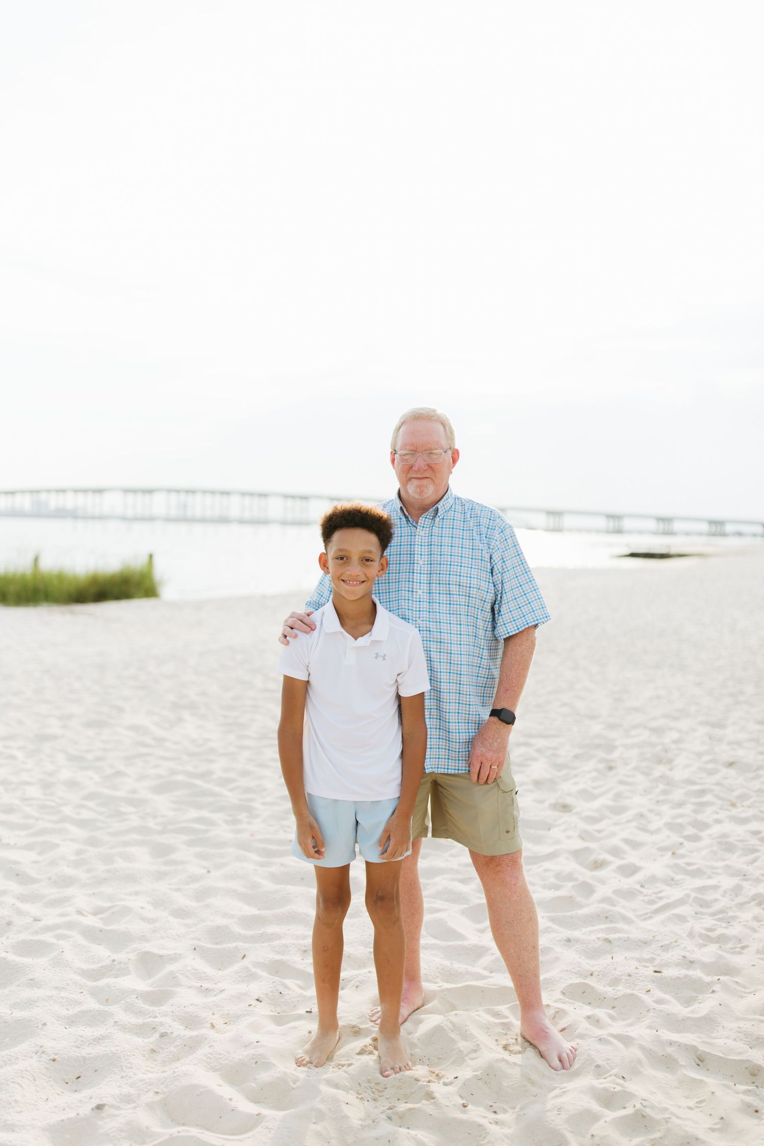 Two people standing together barefoot on a white sandy beach with water and bridge visible in background.