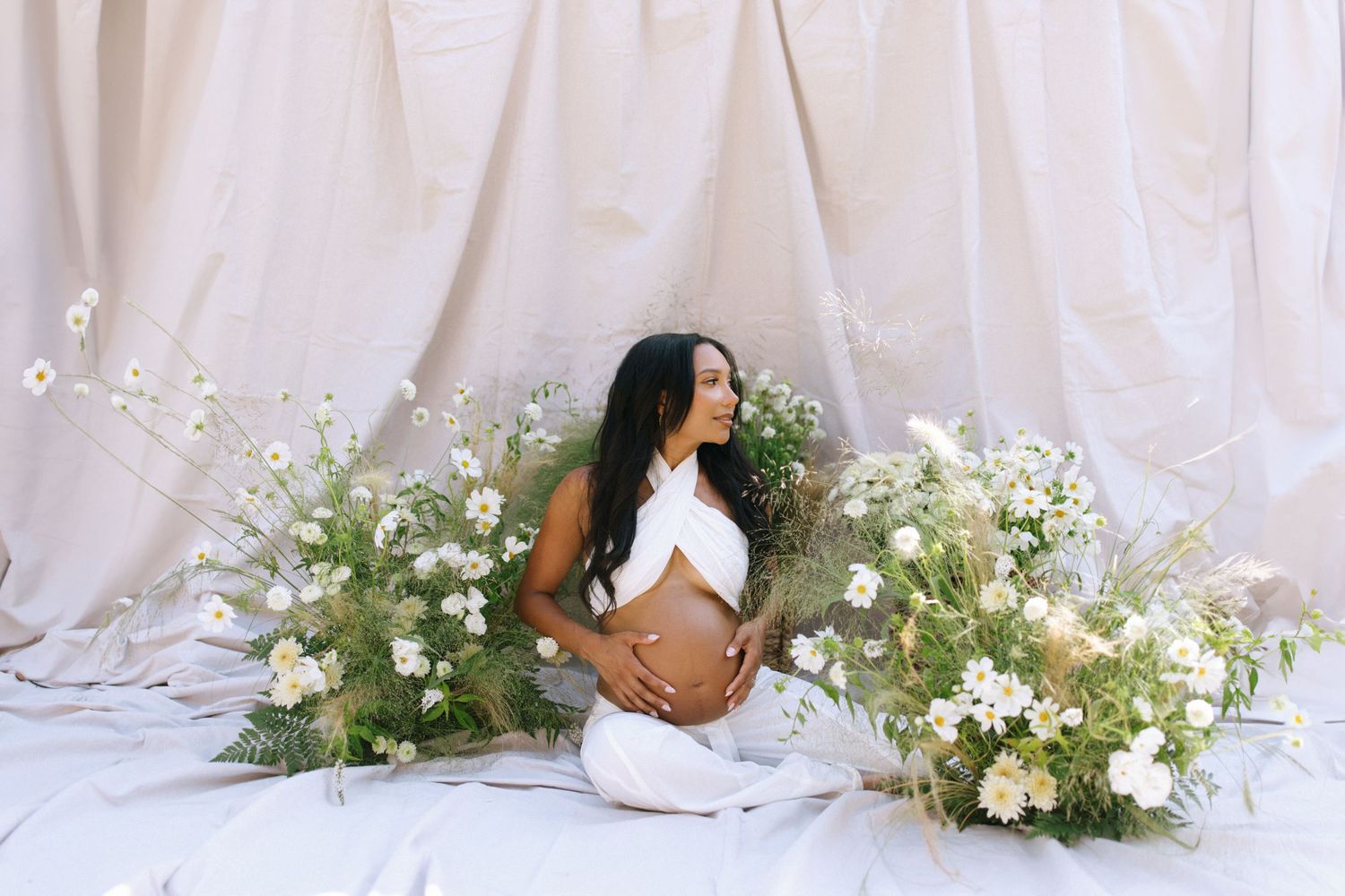 A maternity photo with white flowers and draped fabric creates an ethereal scene on a light backdrop.