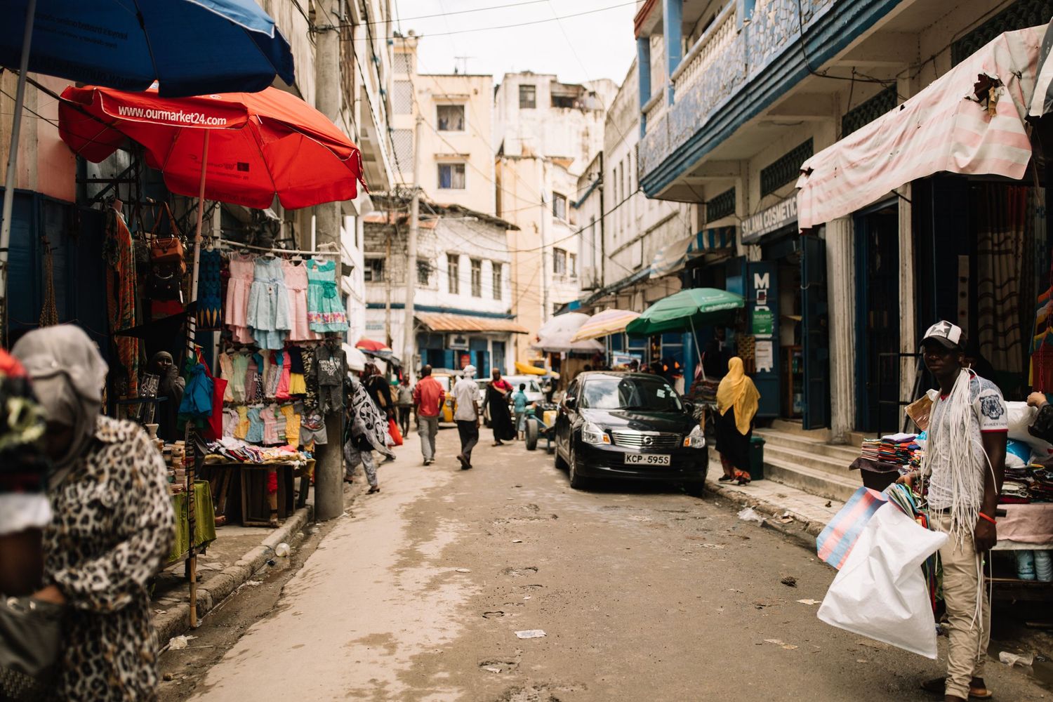 Busy street market in an urban neighborhood with vendors under red umbrellas and parked cars between buildings.