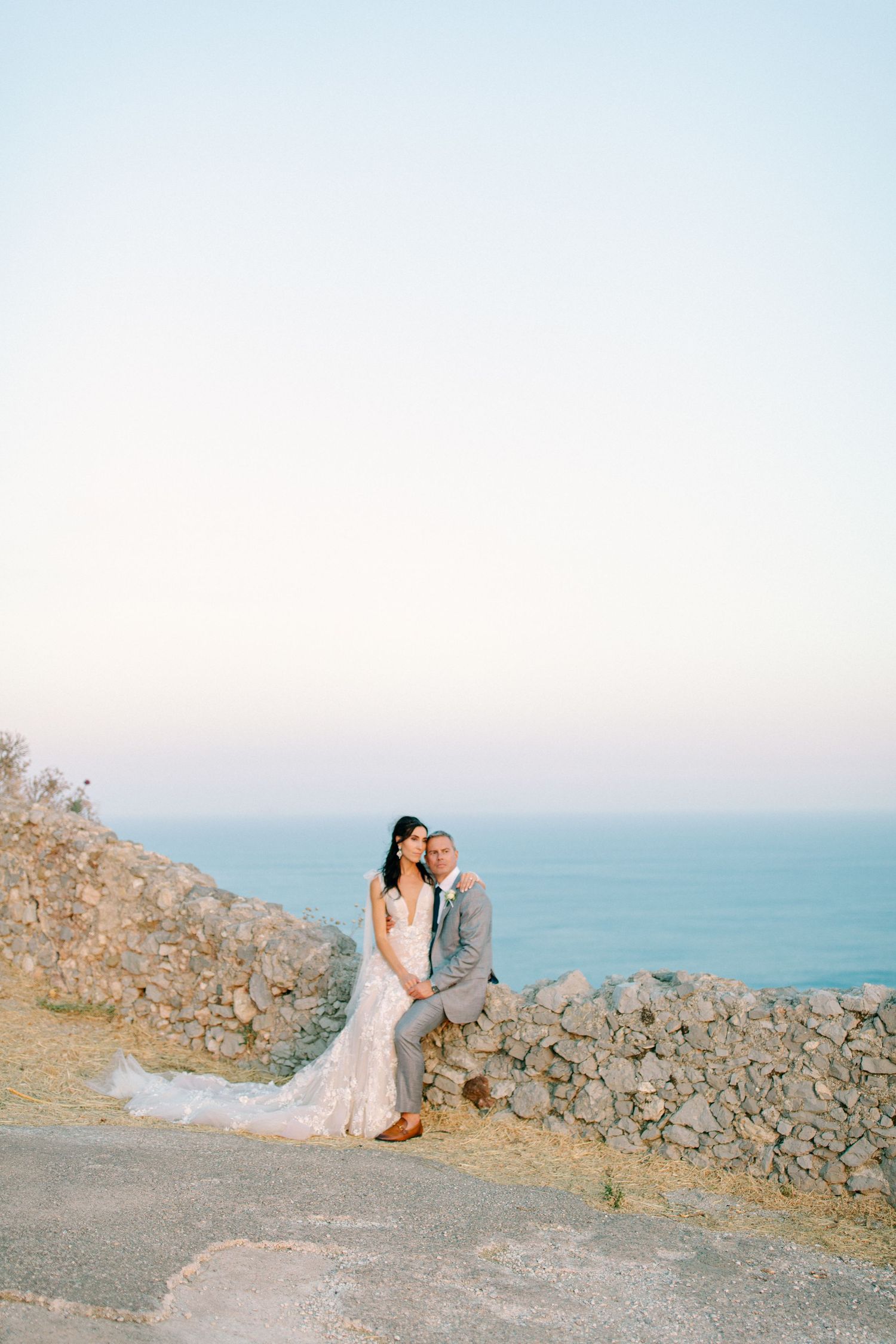A couple in wedding attire stands together on a coastal cliff overlooking the ocean at sunset.