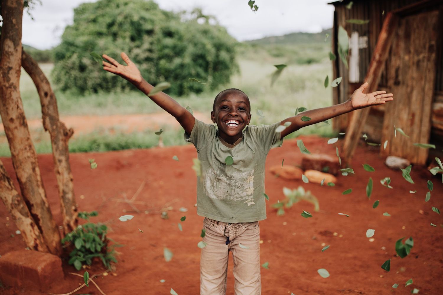 A child jumps joyfully with arms raised outside a rustic building on red clay soil in a rural setting.
