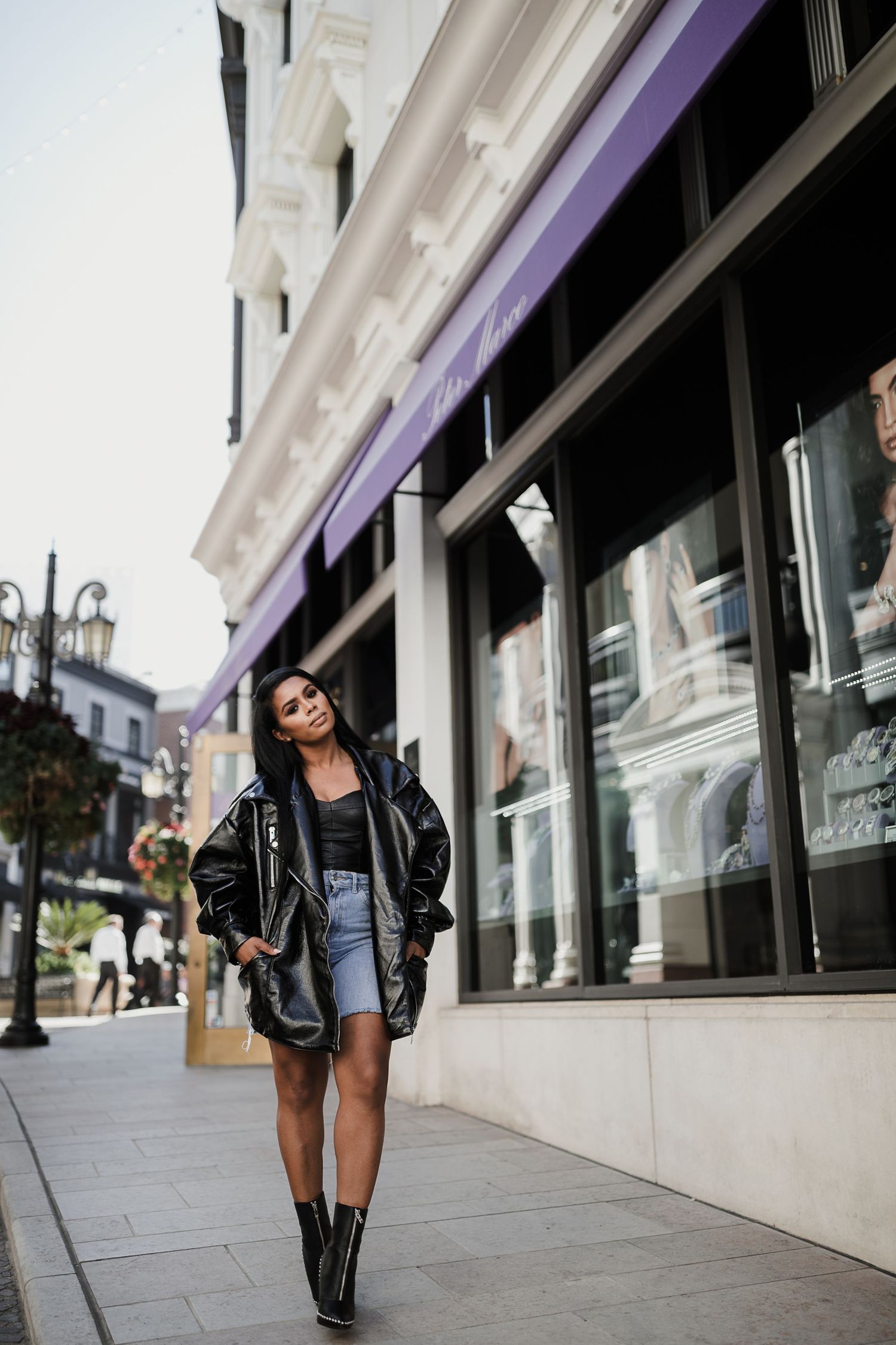 Fashionable woman in leather jacket and denim shorts walking confidently past urban storefronts.