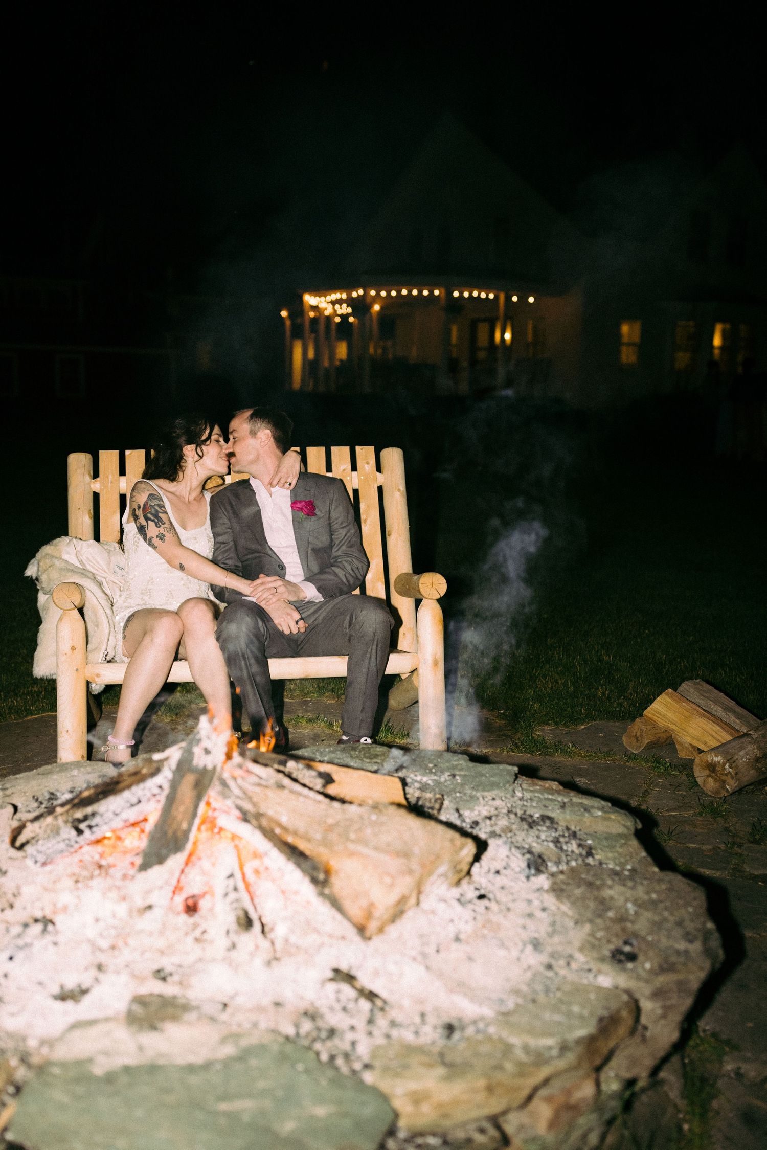 A person relaxes on a rustic log bench near a glowing campfire at night in the wilderness.