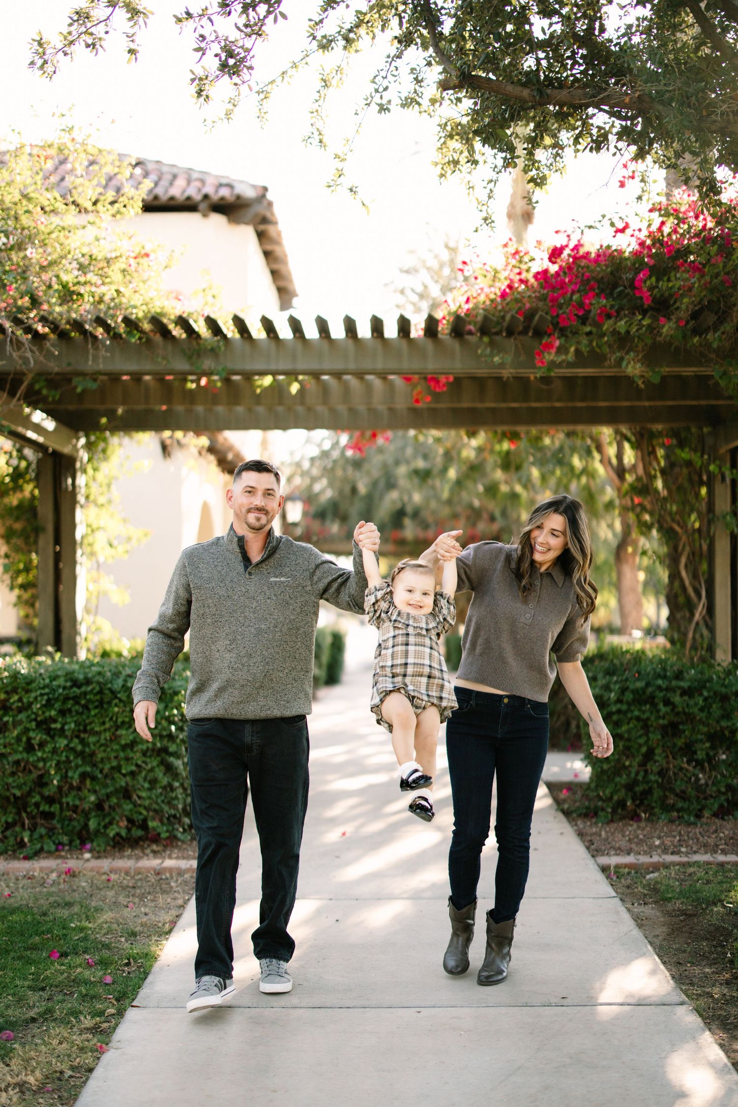 Three people walking together down a sunlit path with trees and pink bougainvillea flowers overhead.