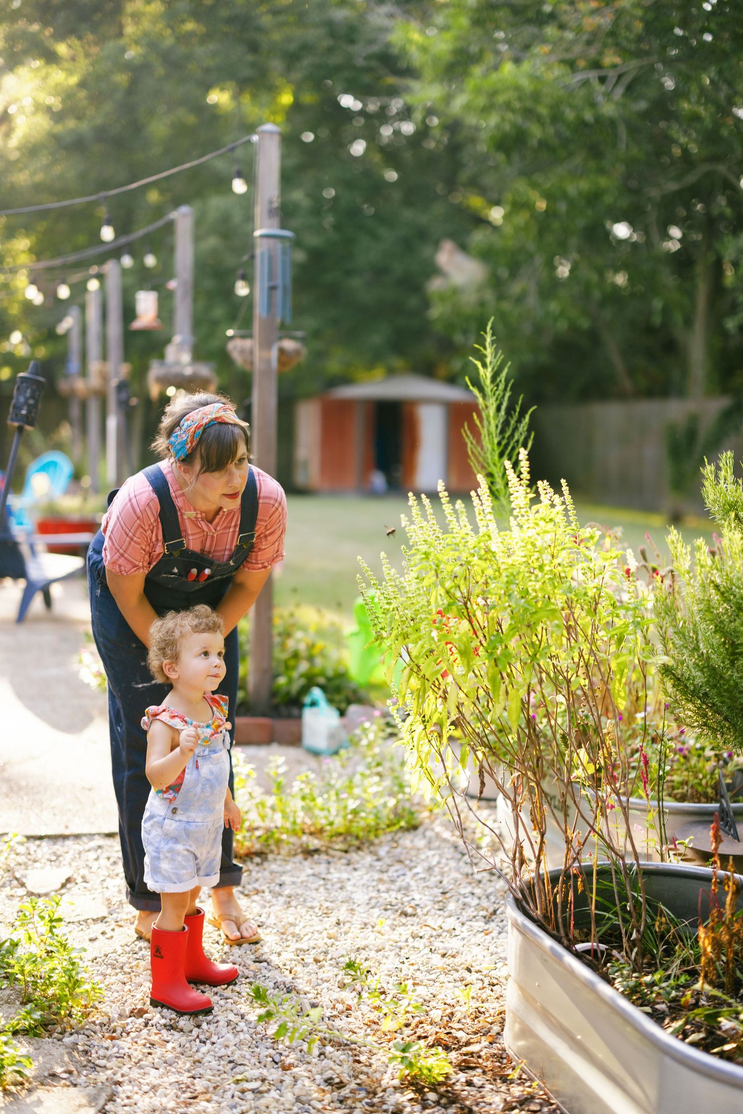 A parent and small child explore a community garden bed filled with herbs and plants on a sunny afternoon.
