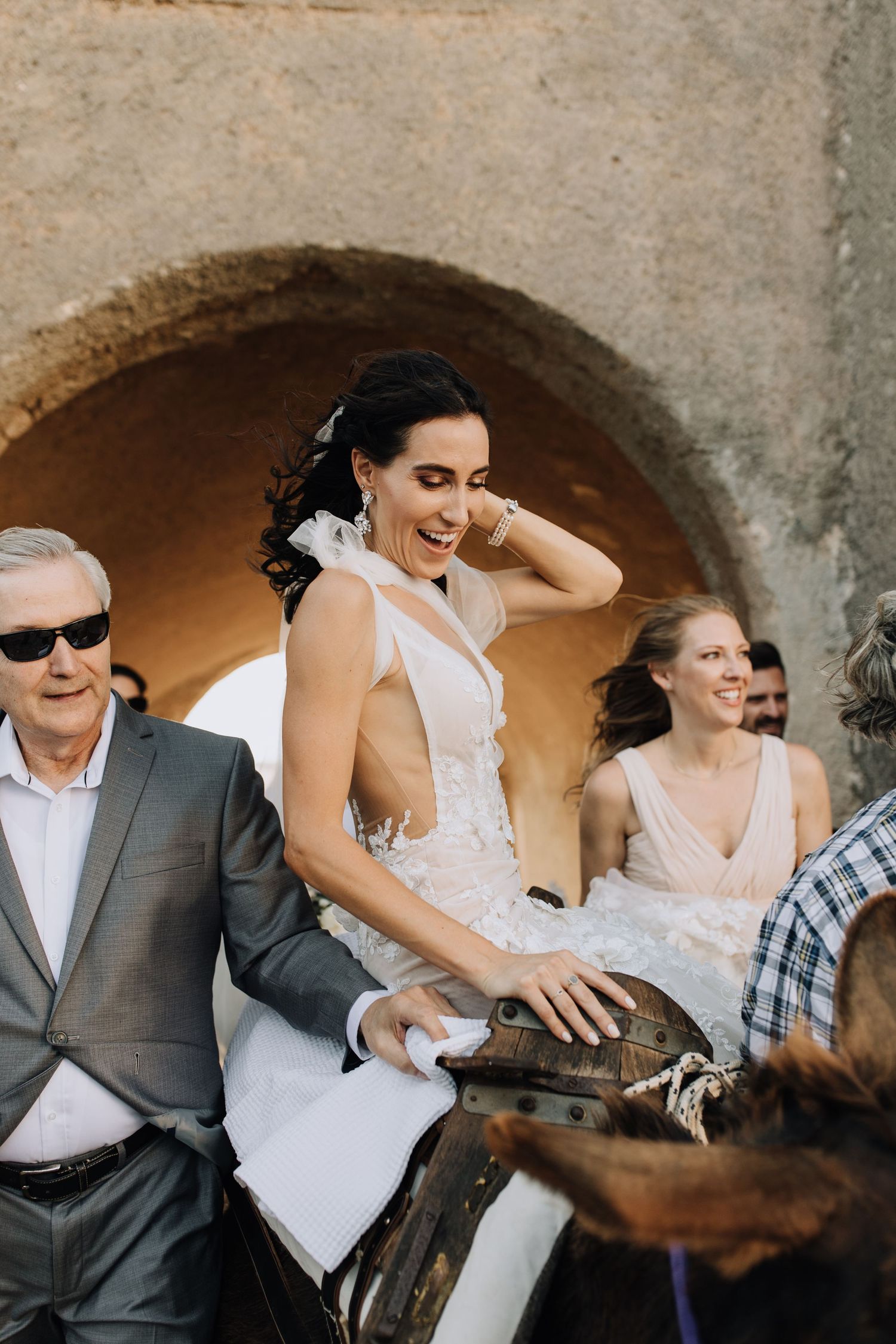 A bride in a white dress laughs joyfully alongside wedding guests under a stone archway.