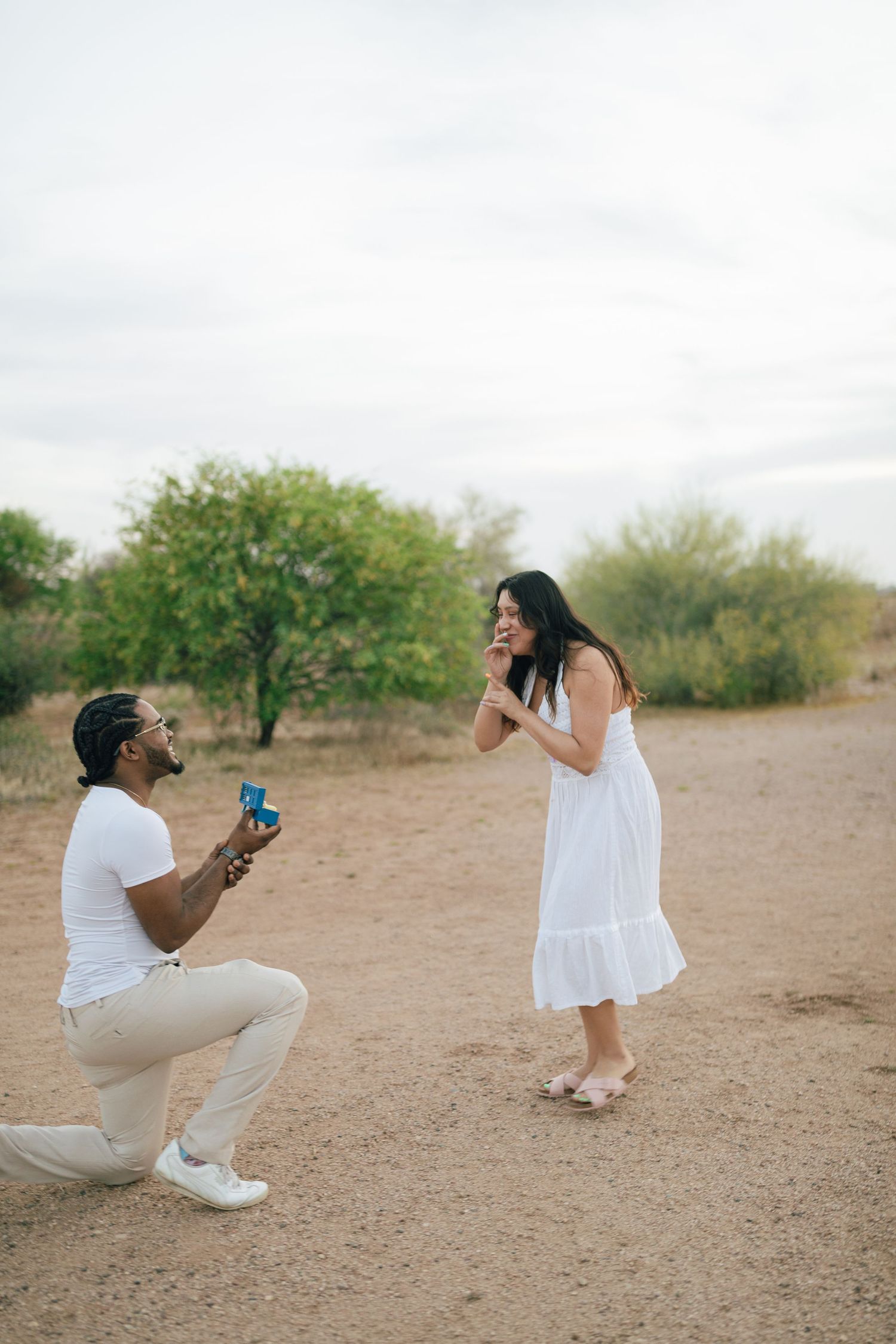 Sweet surprise proposal moment captured in a desert setting with white and tan outfits against natural desert landscape.