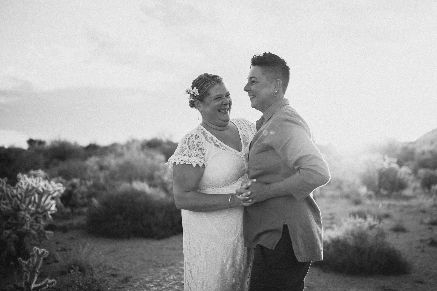 Black and white photograph of a couple embracing outdoors in a scenic mountain landscape setting at sunset.