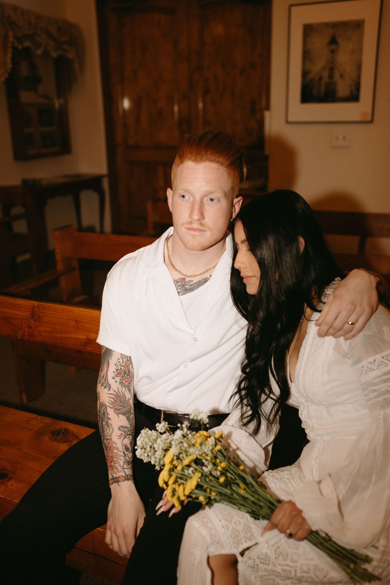 Couple sits together on wooden bench, bride holding bouquet of wildflowers, in warm indoor setting with vintage decor.
