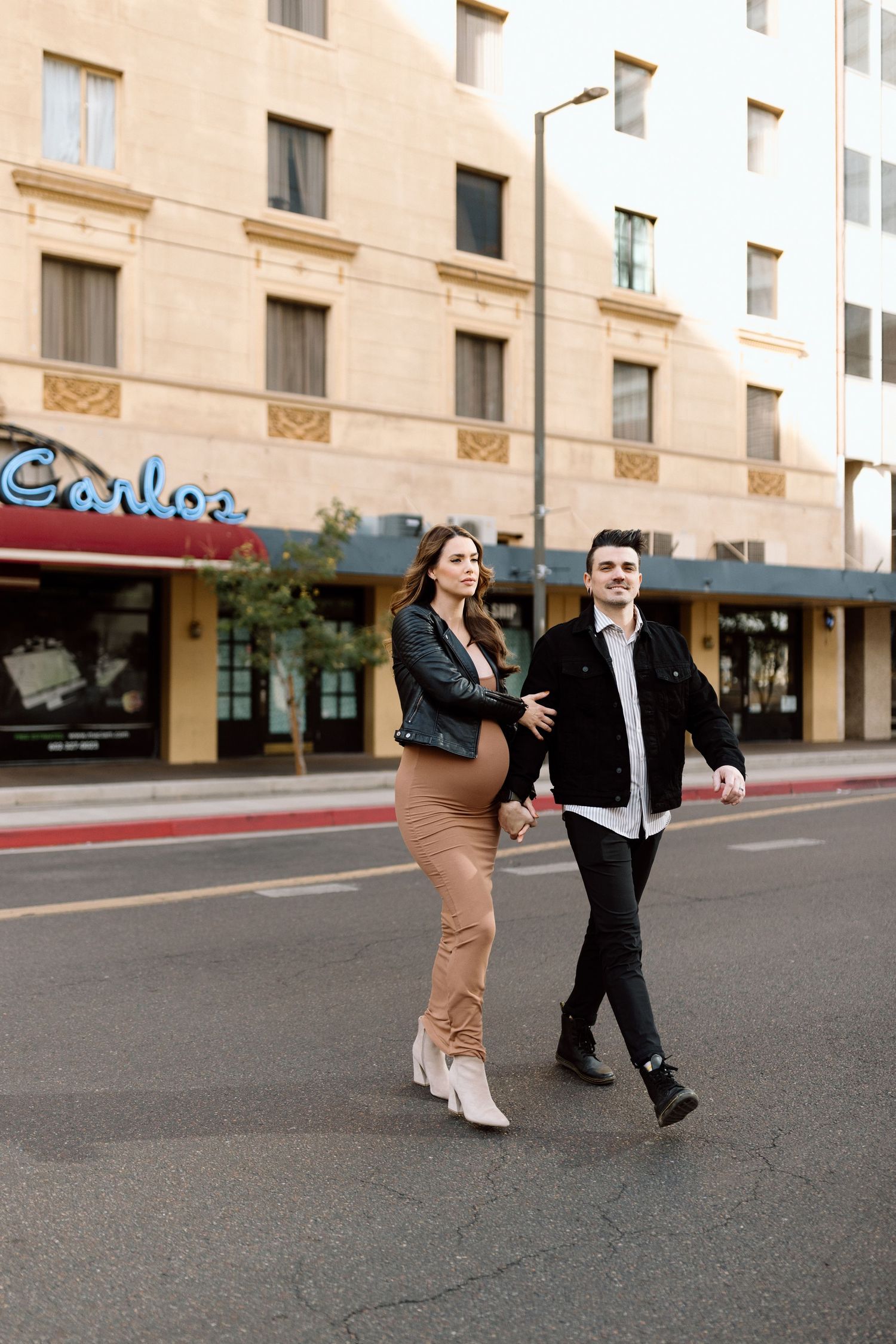 Stylishly dressed couple walks together on a city street in front of beige buildings with storefronts.