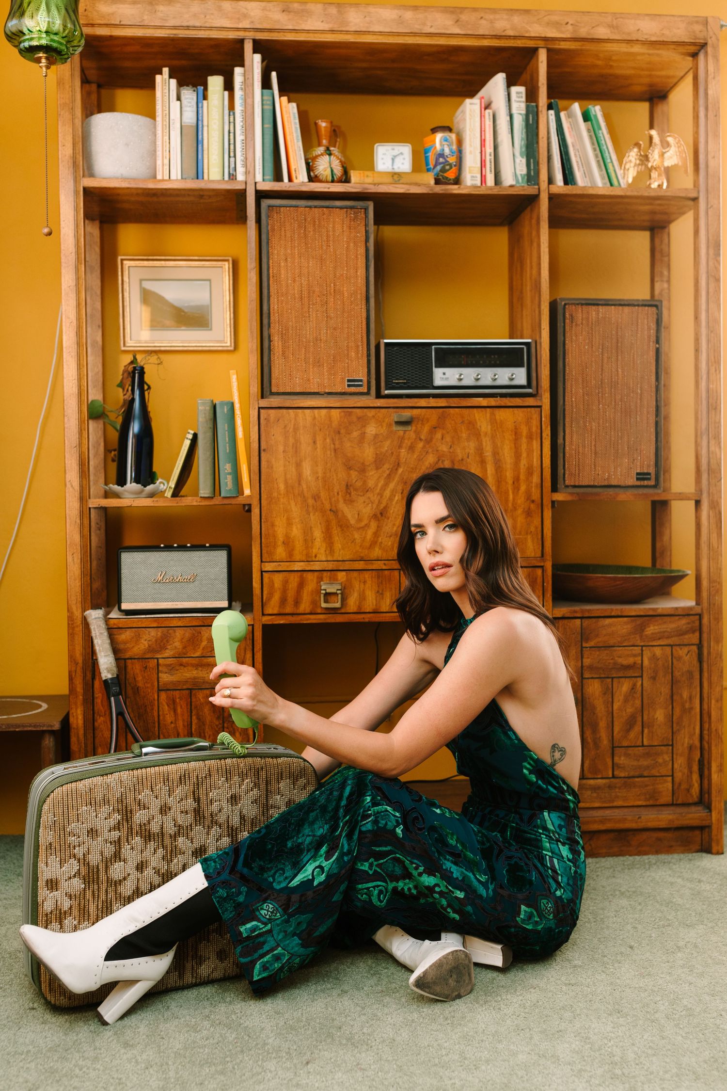 Elegant emerald dress and retro wooden shelving unit create vintage-inspired scene with bottle and stereo components.