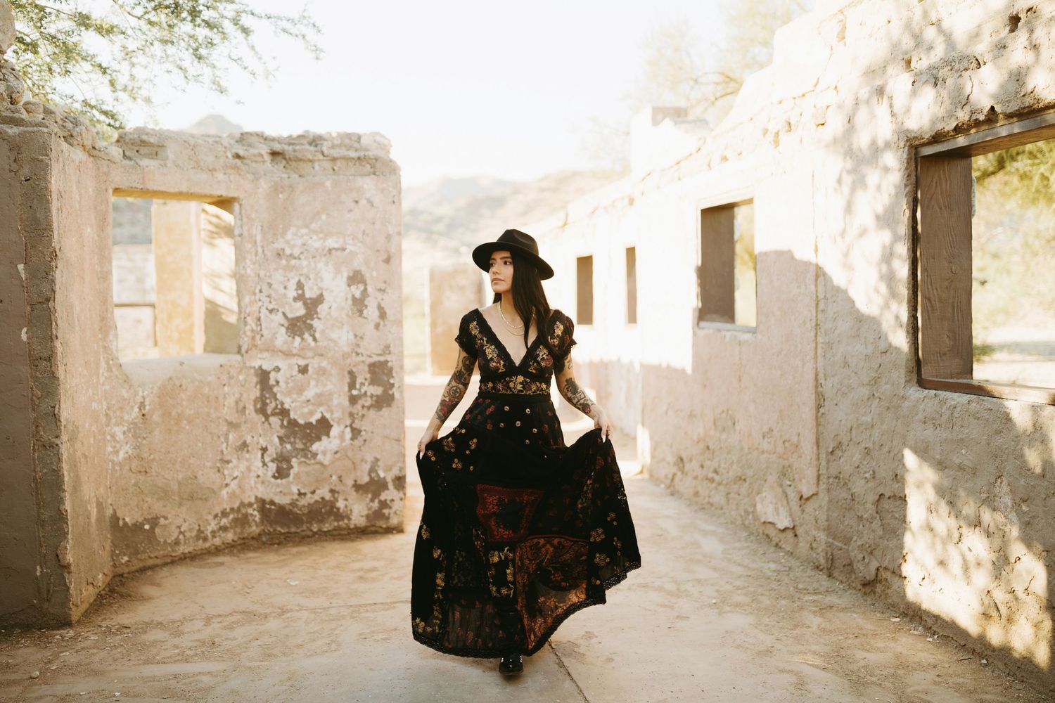 A woman in a flowing black dress and hat poses in a sunlit stone corridor with distressed walls.