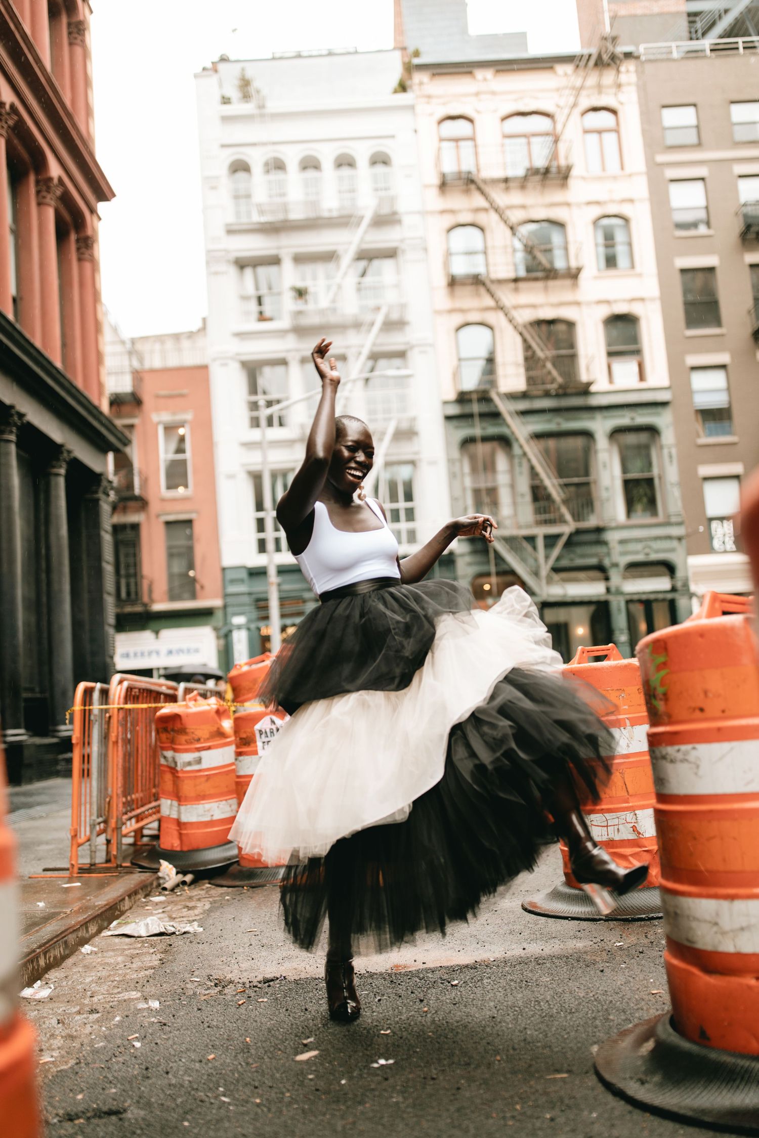Dancer in tulle skirt performs graceful pose on urban street with orange construction barrels in New York City.