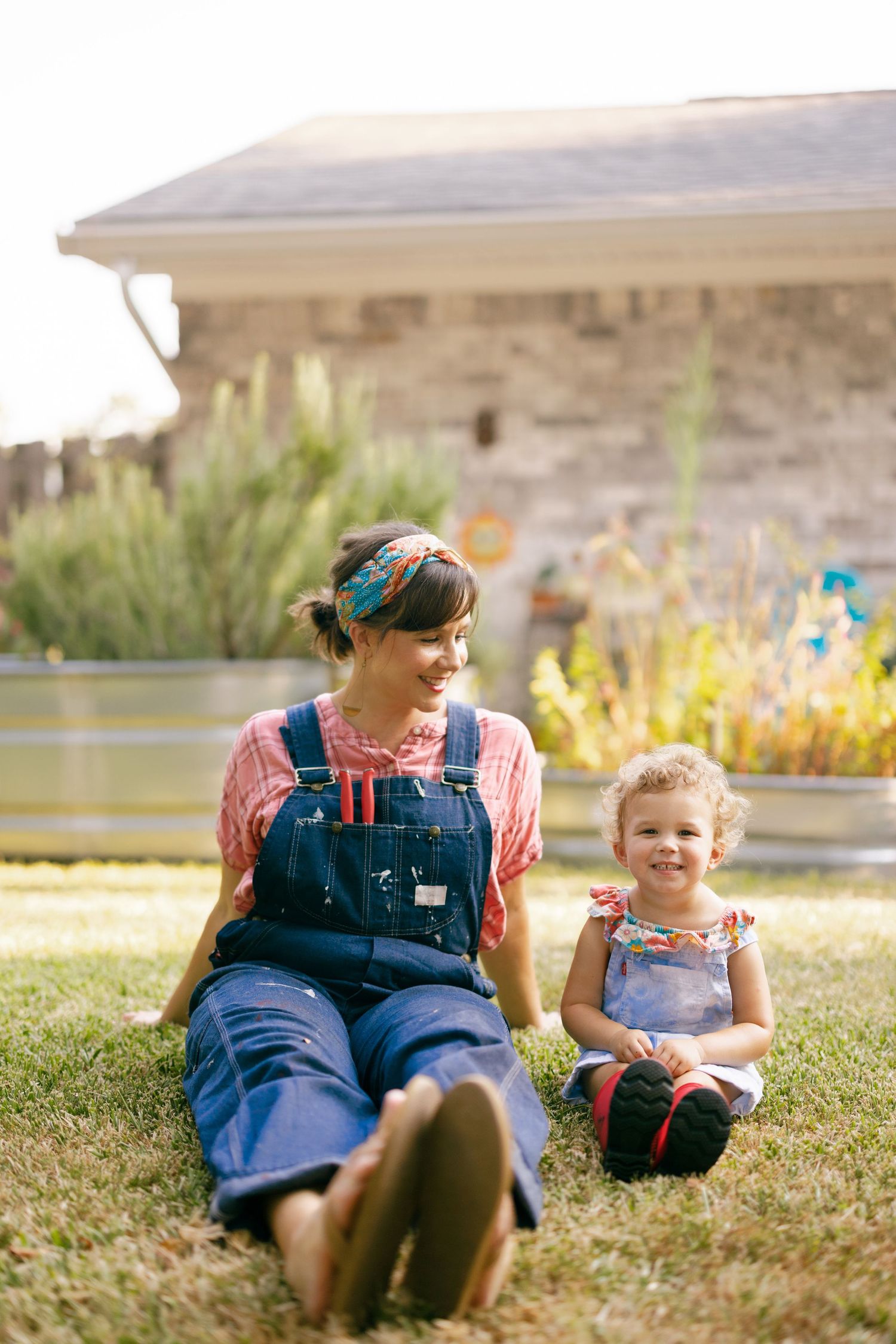 Two people in denim overalls sit together on grass in a garden setting during a sunny afternoon.