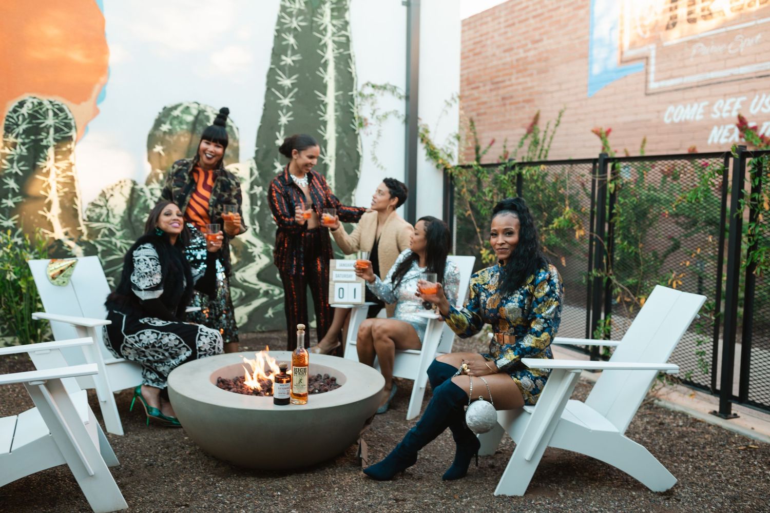 A group gathered around a modern fire pit on an outdoor patio with string lights and brick walls in the background.