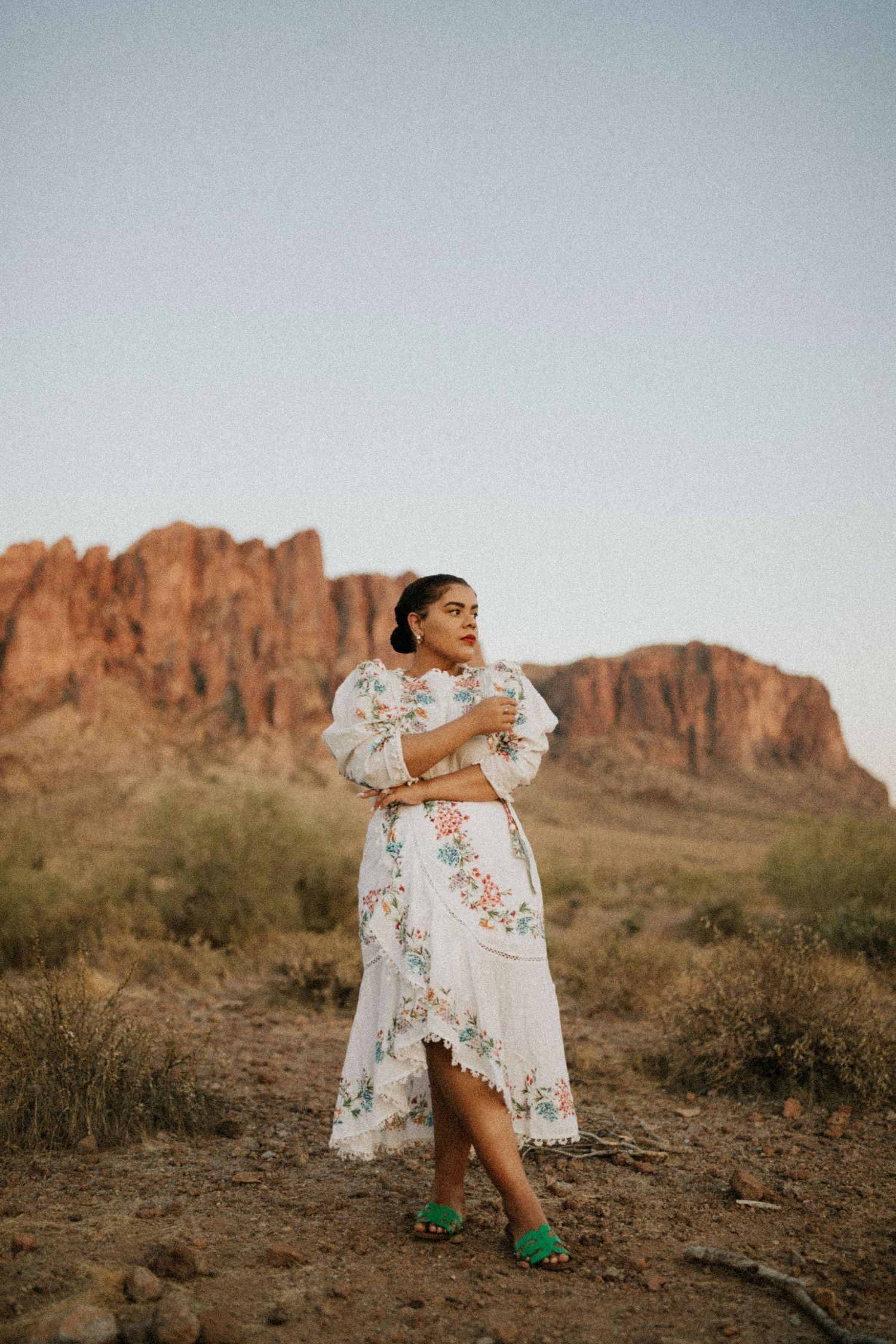 Person in a white floral dress poses in front of dramatic desert mountains at sunset against rugged desert landscape.