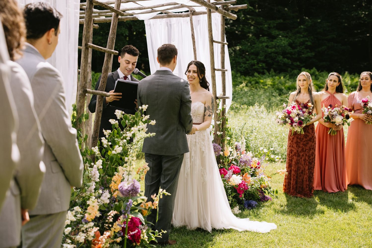 An outdoor garden wedding ceremony with attendees wearing warm colors and a floral-decorated arch in a natural setting.