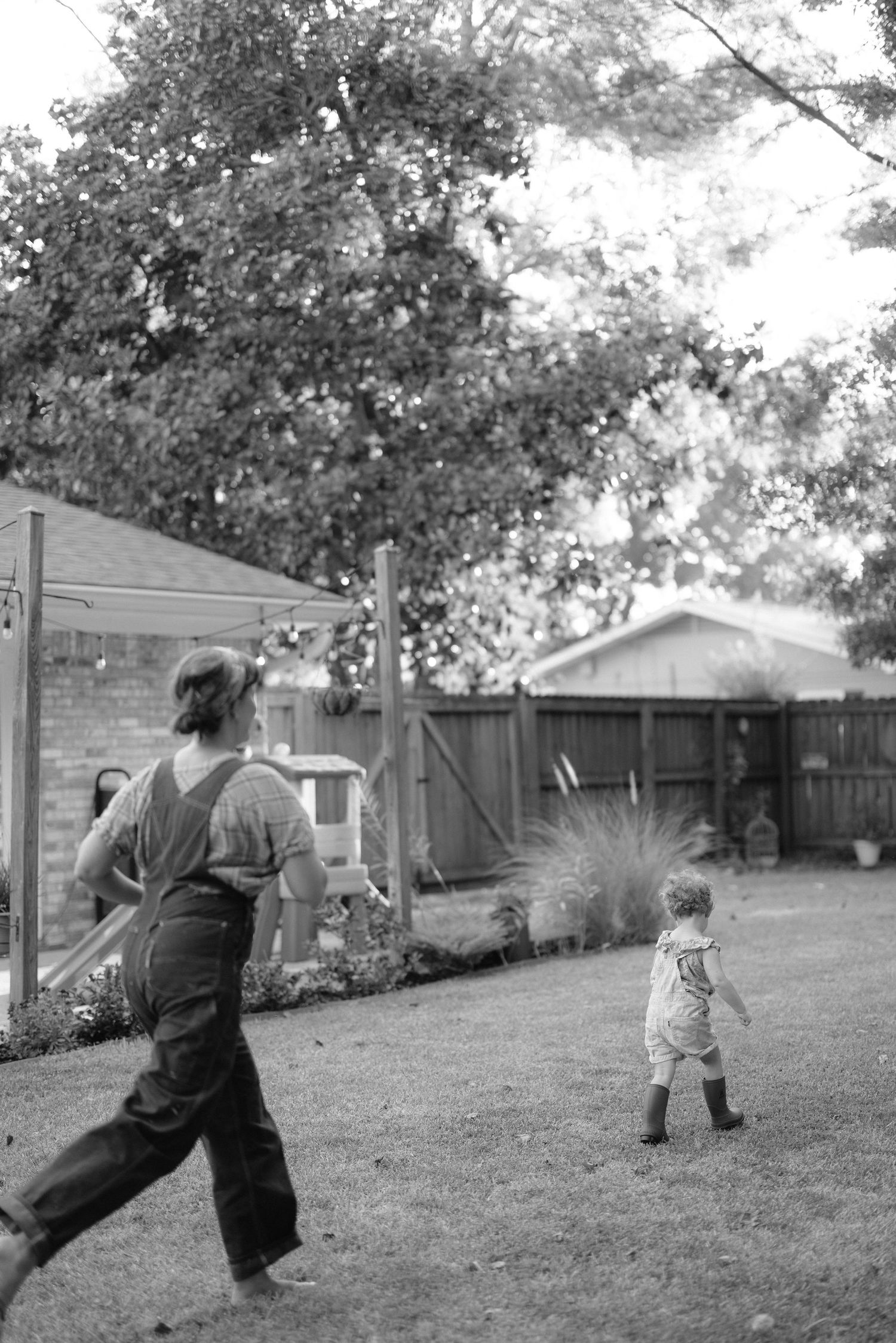 Black and white photo of two people playing together in a backyard with trees and a fence in the background.