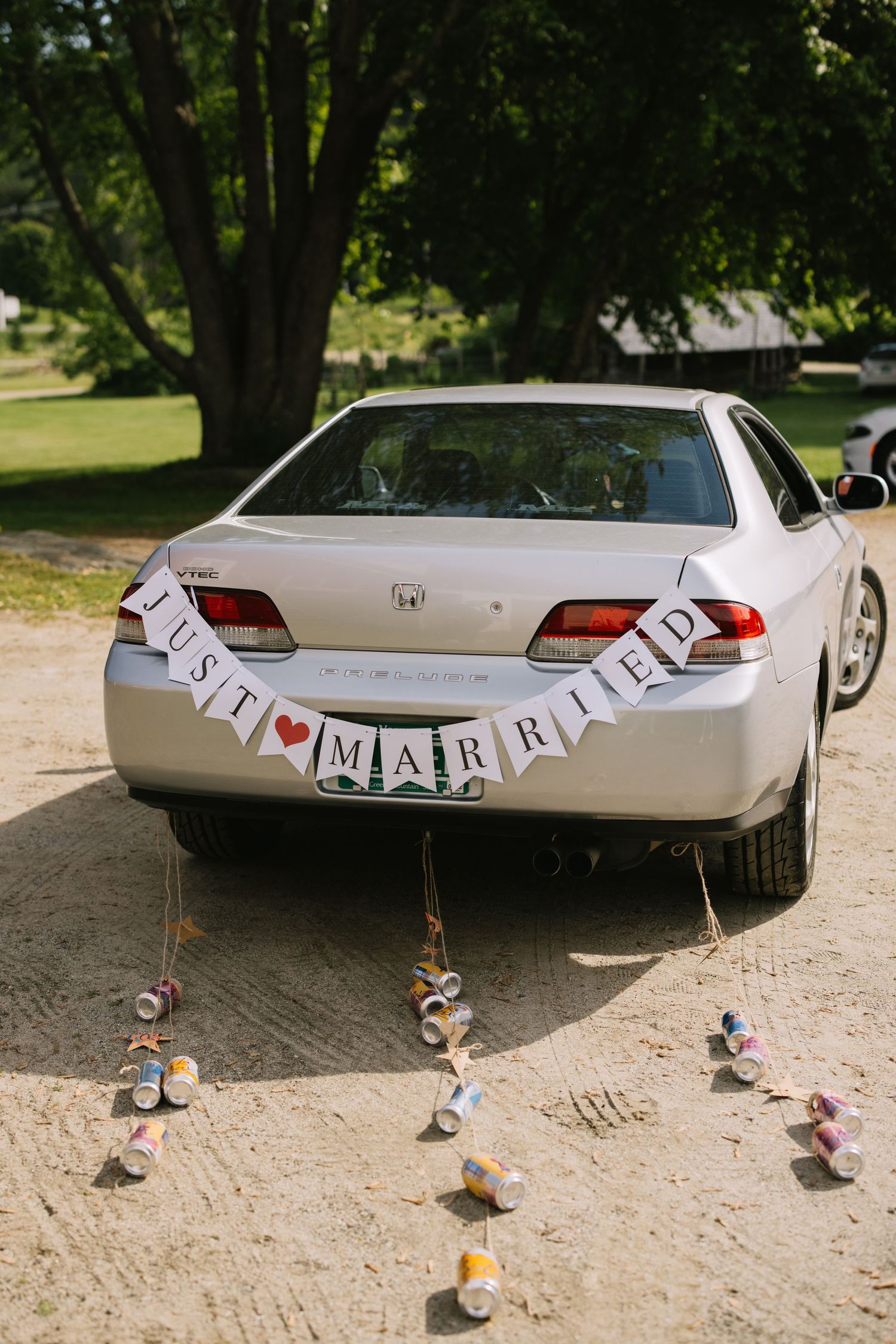 A silver Honda car decorated with 'Just Married' banner and tin cans tied to the back bumper parked under trees.
