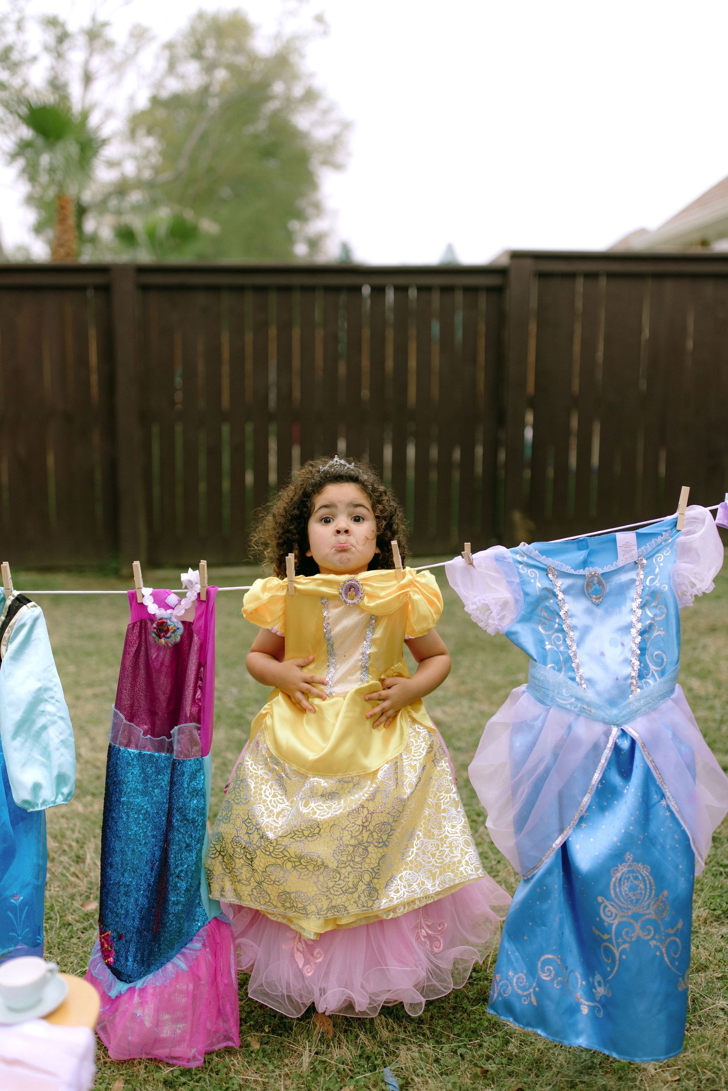 Colorful Disney princess dresses hanging on a clothesline in a backyard with a wooden fence backdrop.