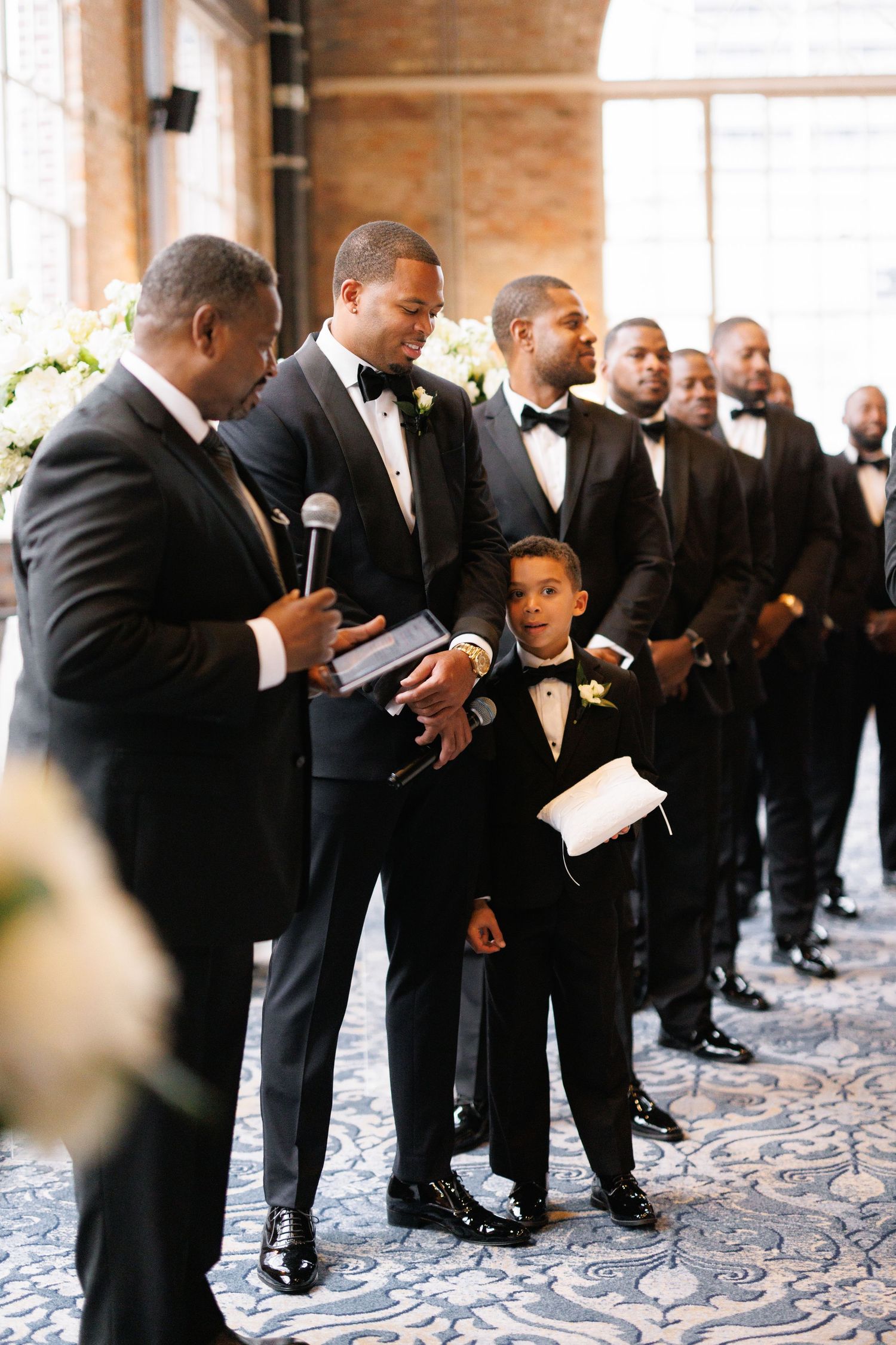 A young ring bearer in a tuxedo stands among groomsmen during a formal wedding ceremony.