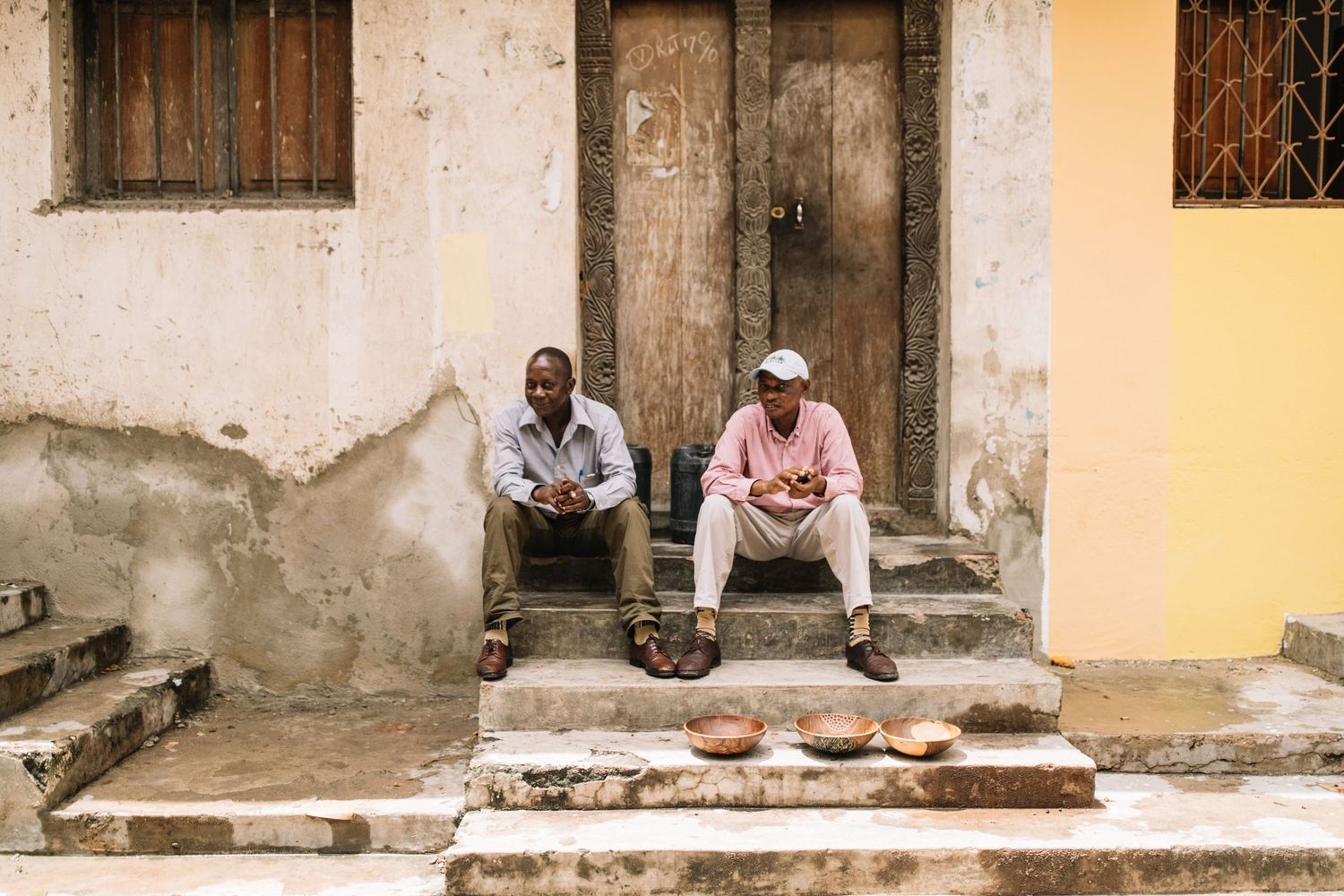 Two people sit on stone steps outside an old weathered doorway of a building with peeling paint and a yellow wall.