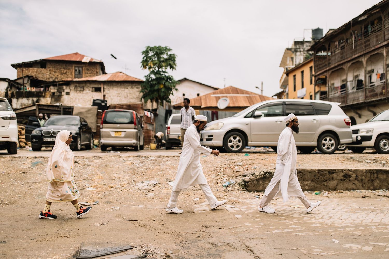 Three people in white protective suits walk in single file past parked cars on a dirt road in an urban area.