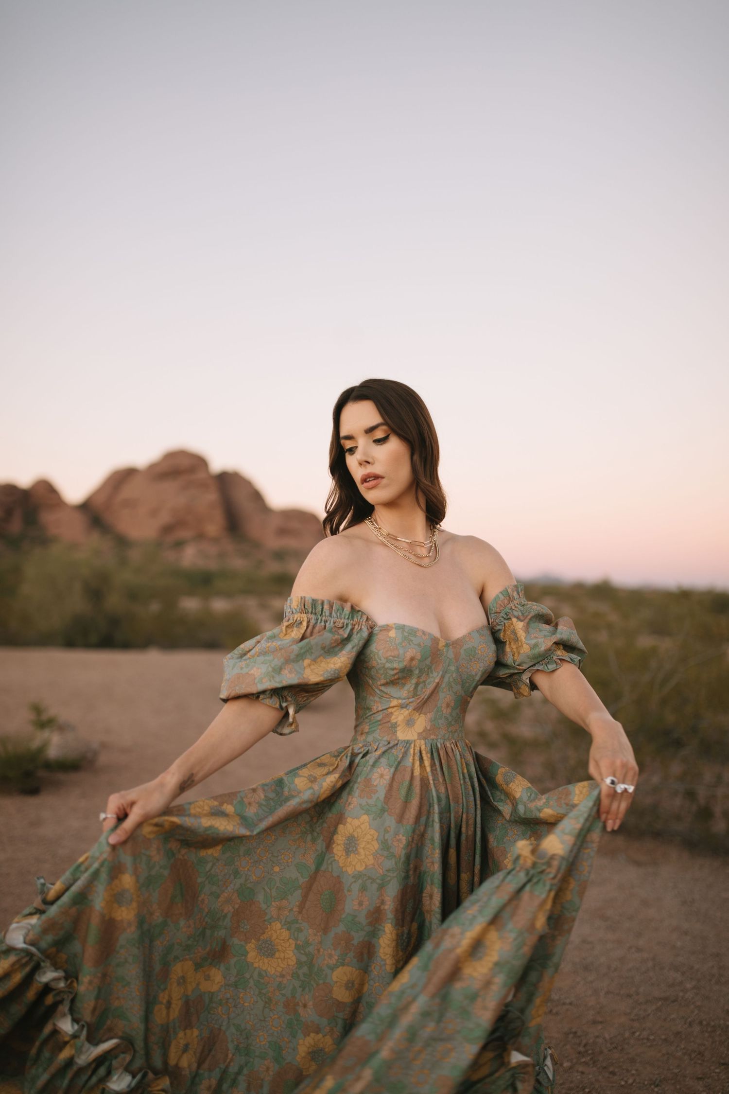 A person wearing a floral empire waist dress poses gracefully in a desert landscape at sunset.