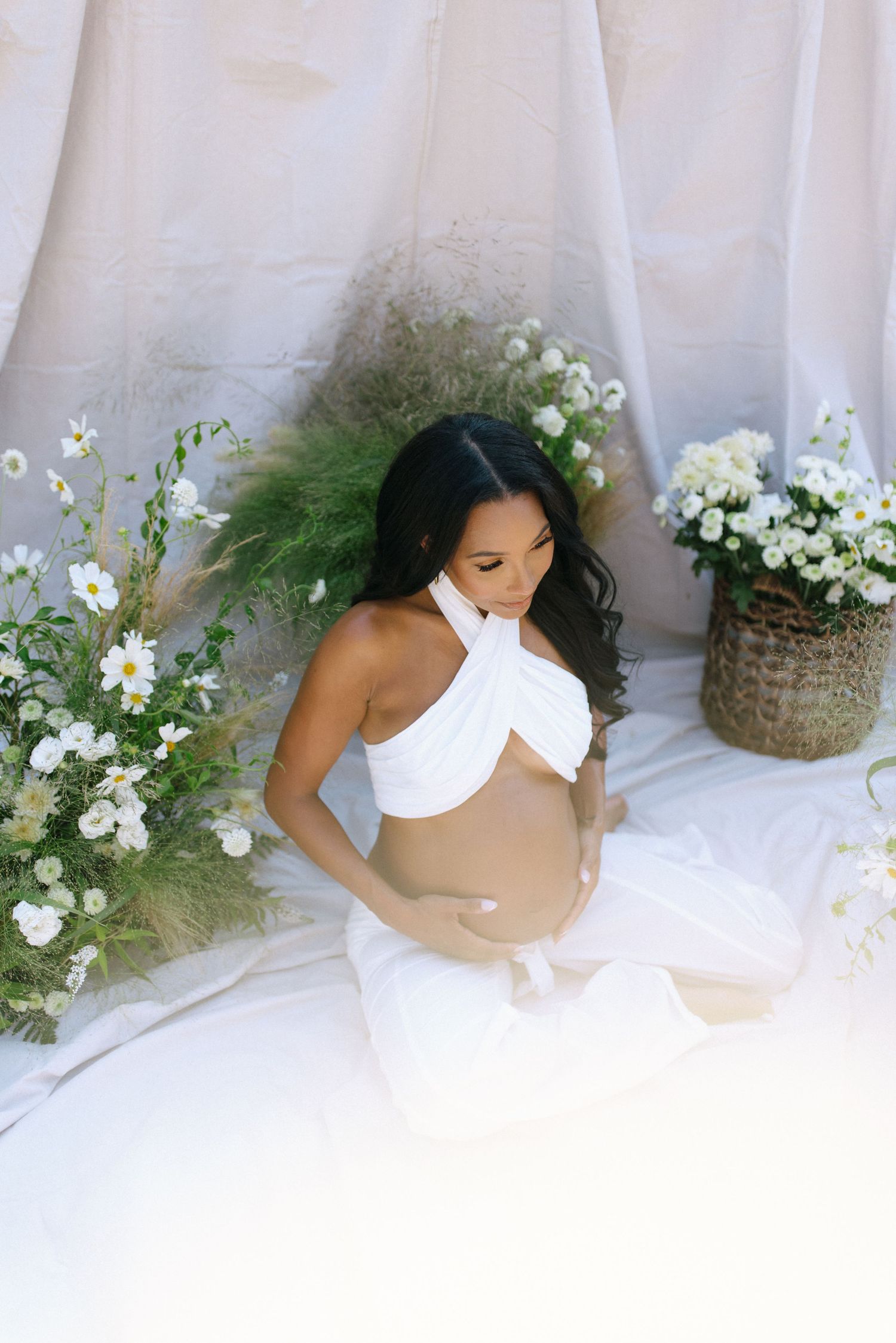 Ethereal maternity photo with white floral arrangements and greenery surrounding figure in draped white fabric on light backdrop.