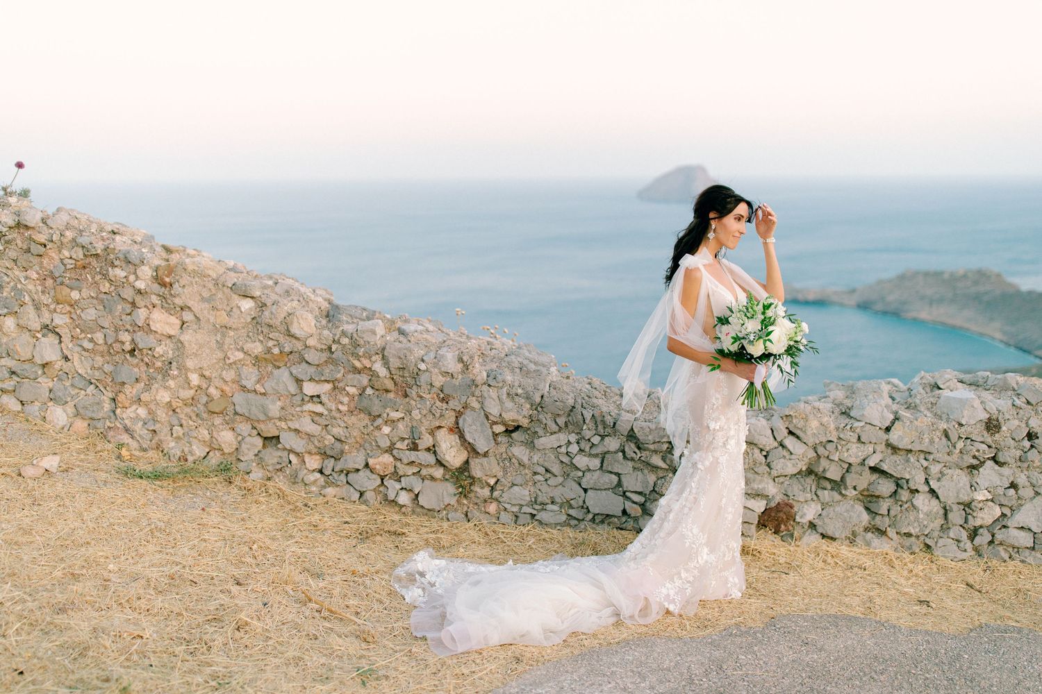 A bride in a lace wedding dress holds a blue and white bouquet on a rocky cliff overlooking the ocean.