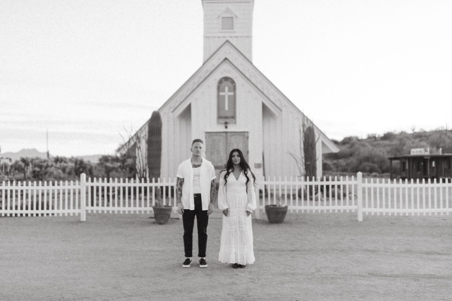 Black and white photo of a couple standing in front of a white country church with a picket fence.