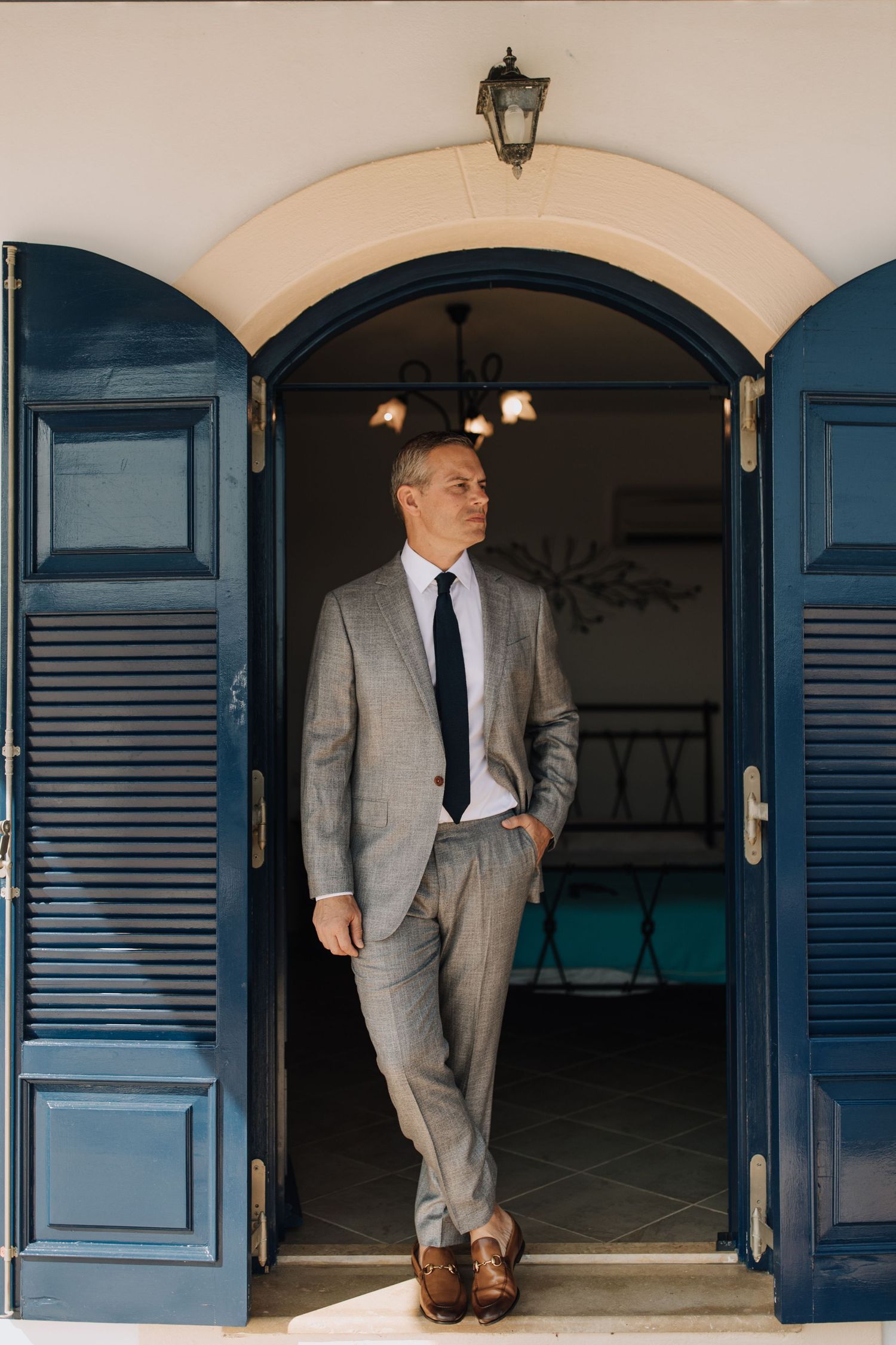 A person in a gray suit and tie stands casually in a doorway with blue shutters and an arched entryway.