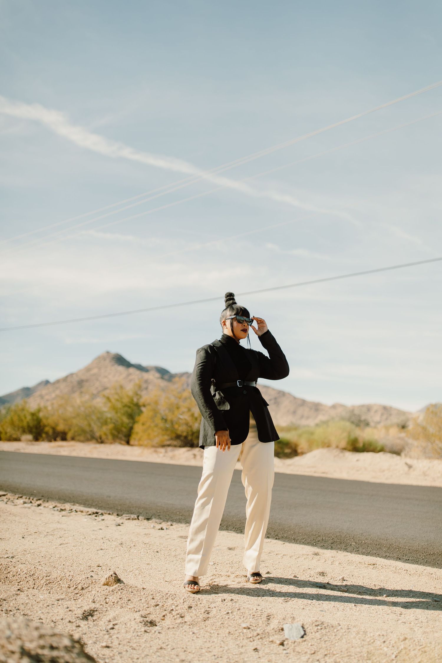 Person in black and white attire walking along a desert road with mountains in the distance against blue sky.