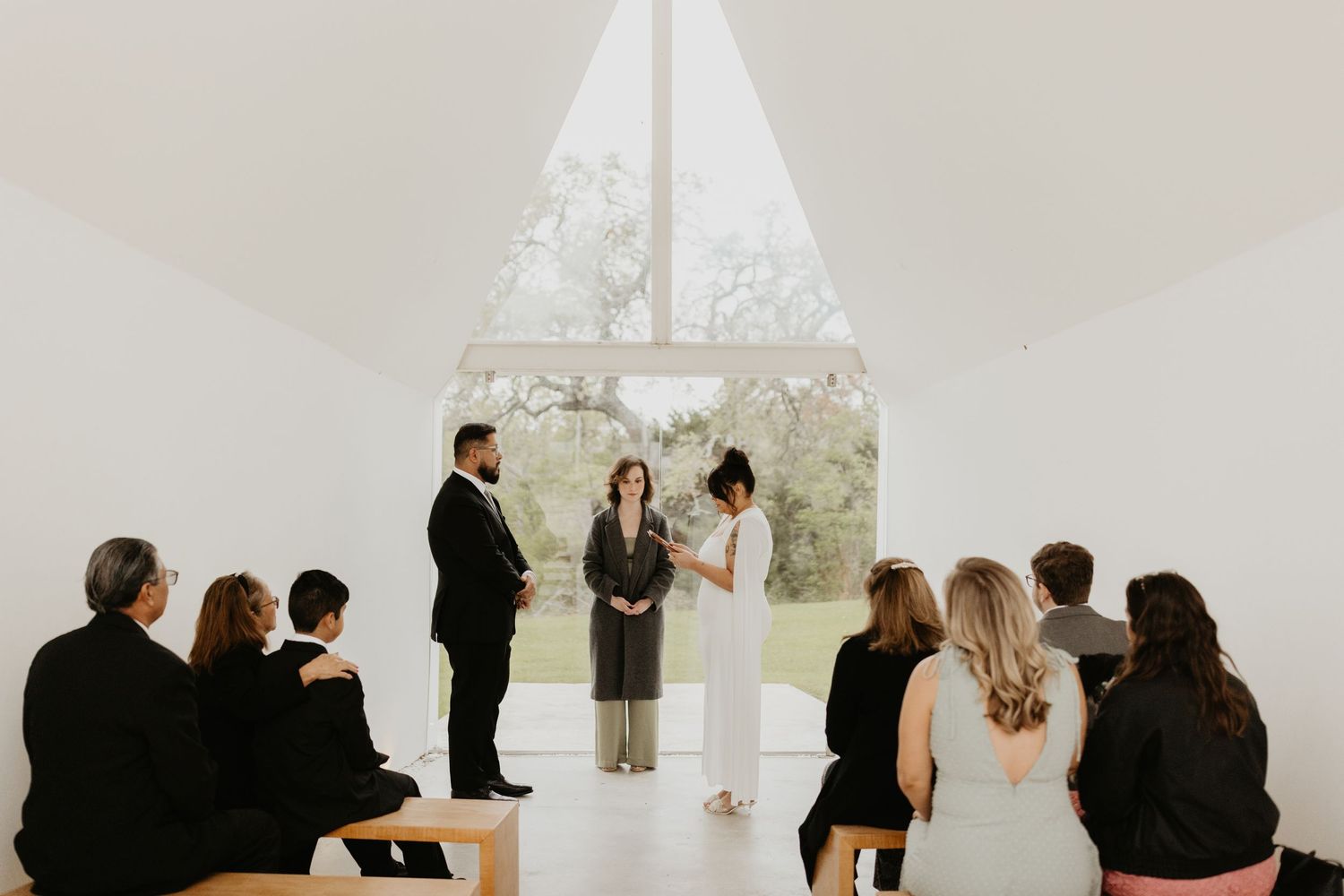 An intimate indoor wedding ceremony takes place in a modern white chapel with floor-to-ceiling windows overlooking nature.