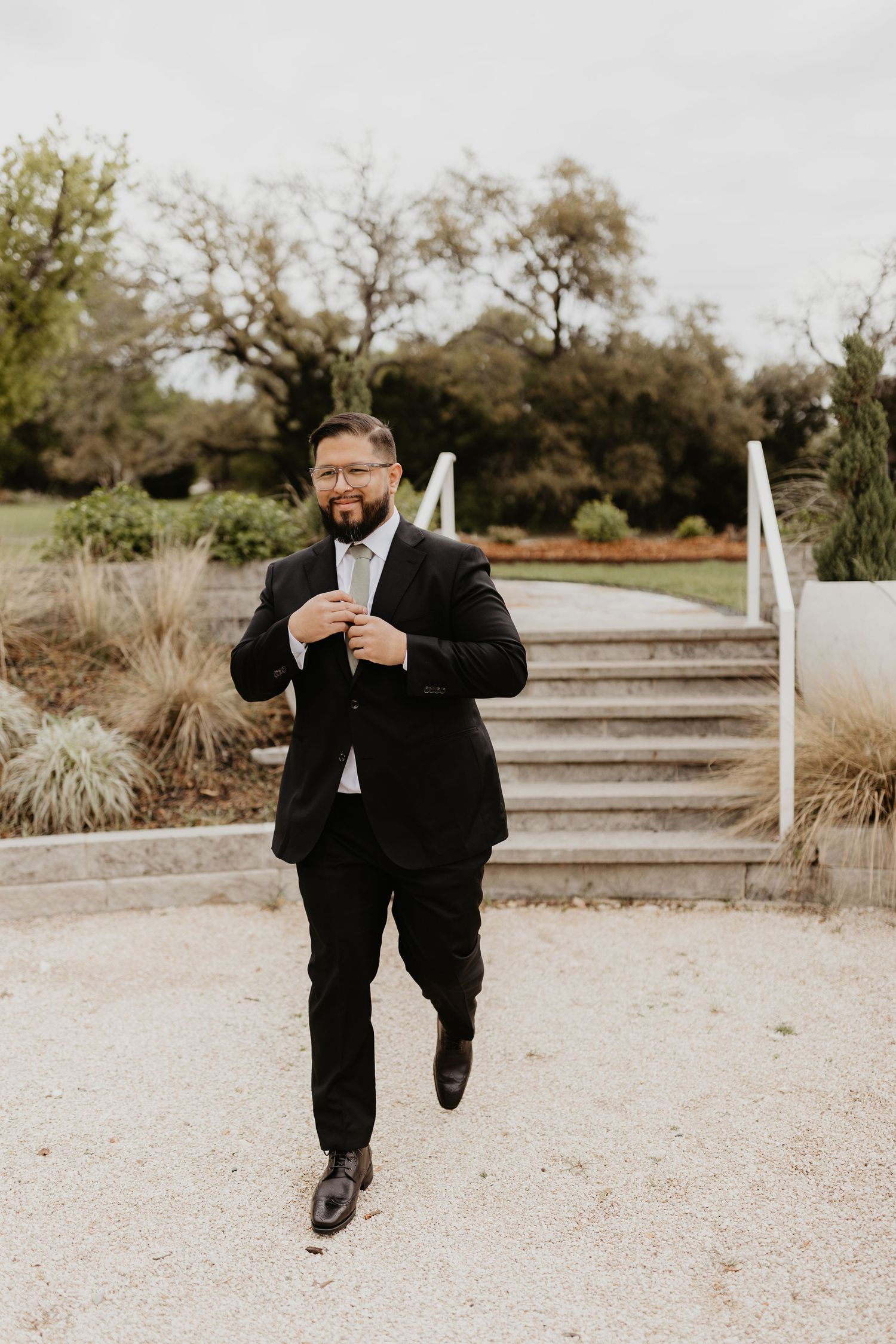 A person wearing a black suit and walking outdoors in a garden setting with stone steps and ornamental grasses.