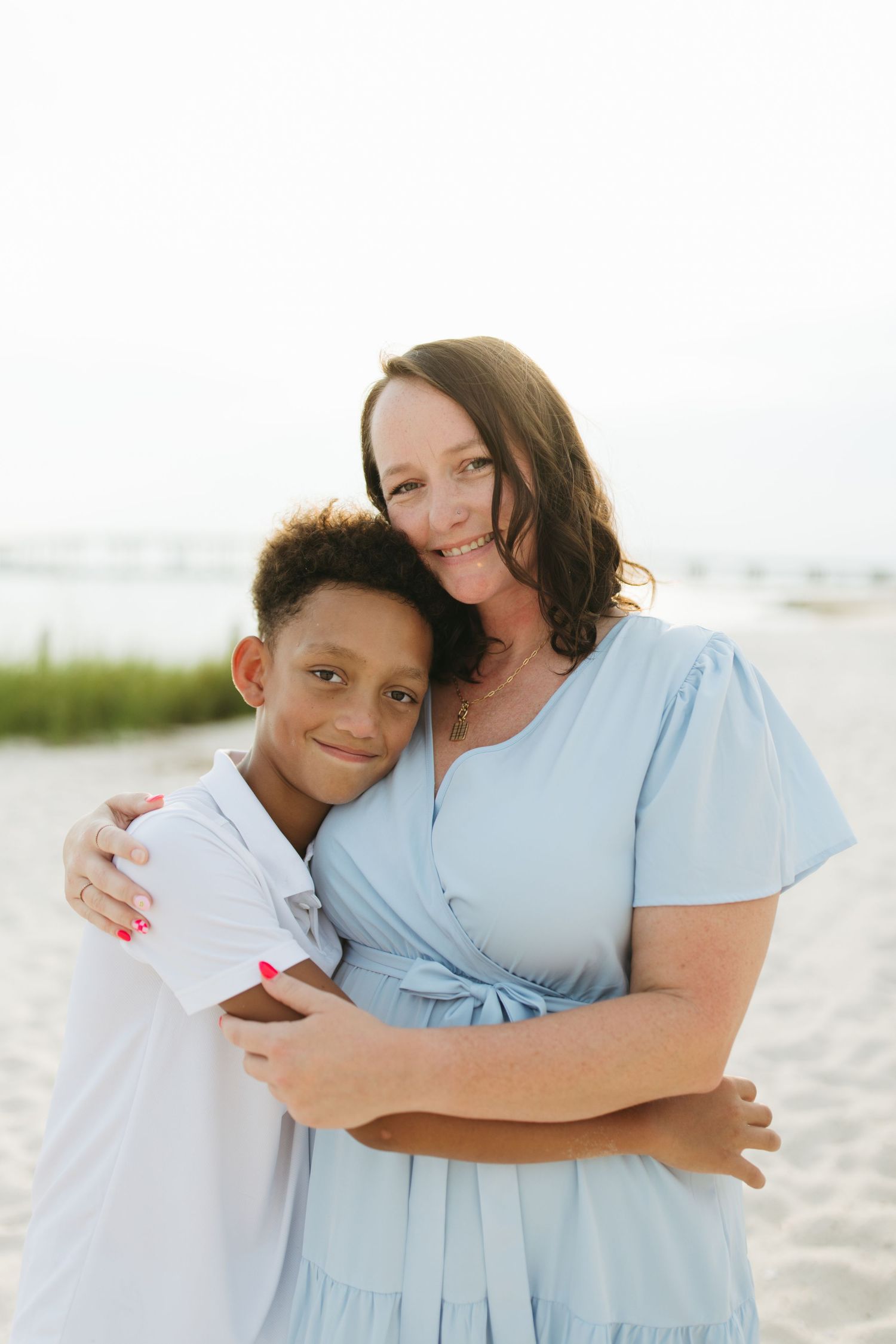 Mother and child embracing while wearing coordinated blue and white outfits on a sunny beach.