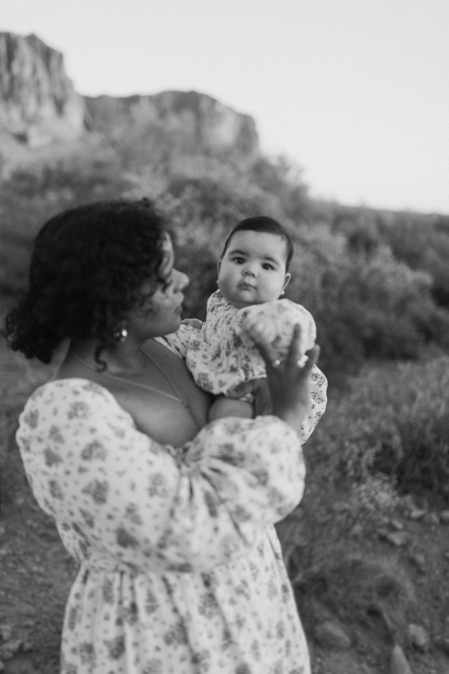 A tender black and white photograph shows a woman in a floral dress holding a baby against a mountain backdrop.