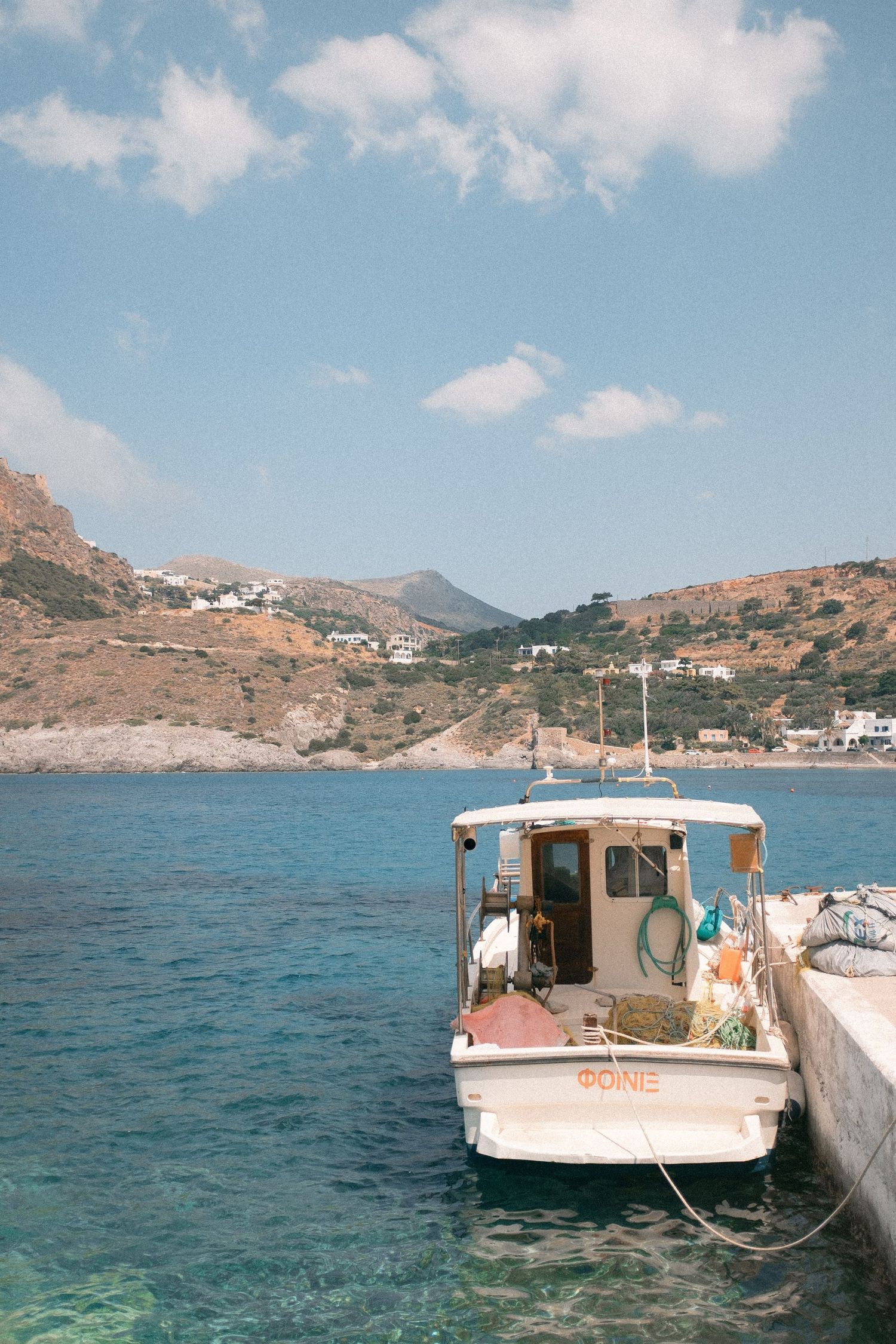 Scenic Mediterranean coastline with moored fishing boats against blue waters and rugged mountains under puffy clouds.