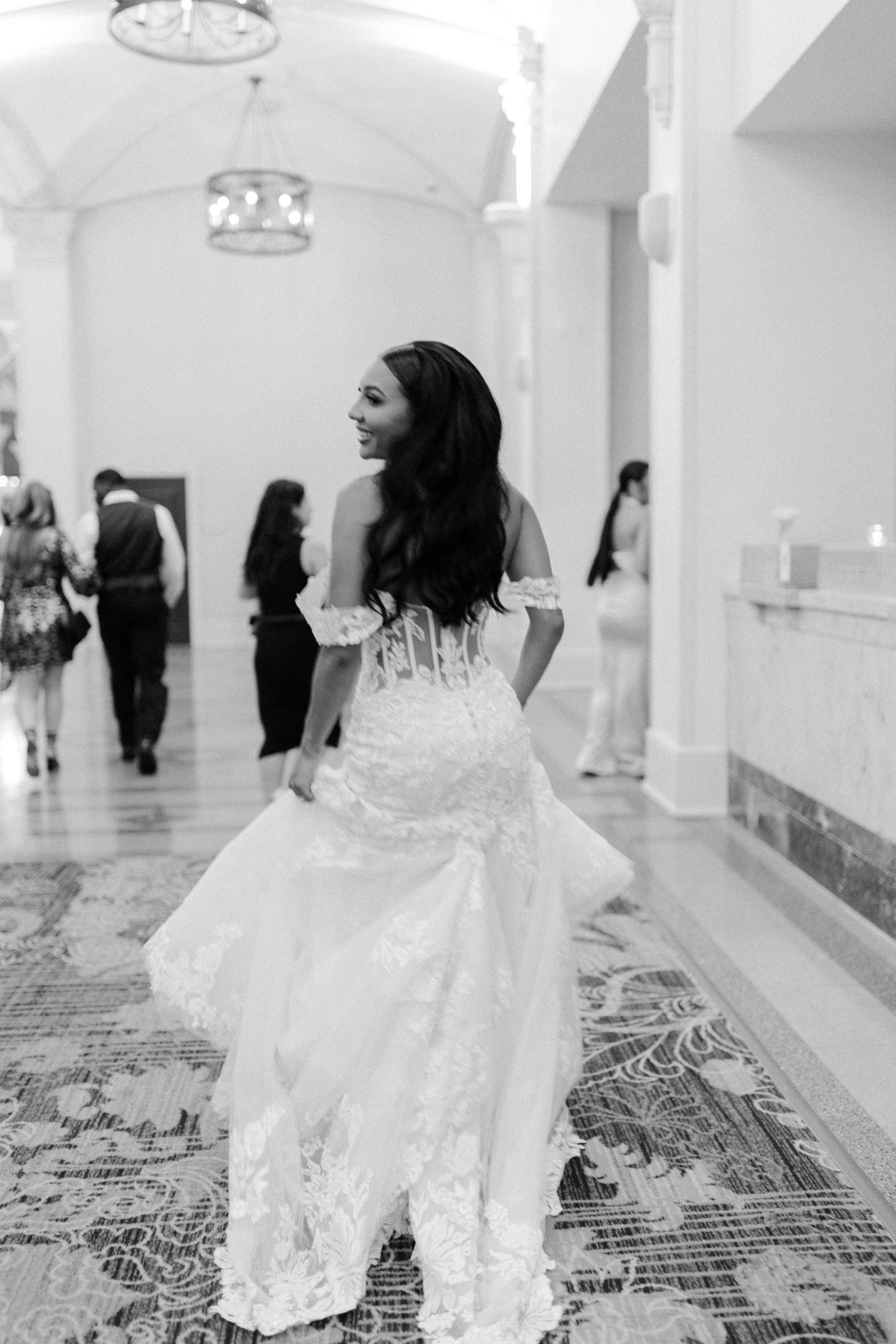 A person in a flowing white wedding dress walks through an ornate mosque corridor with patterned carpet floors and white pillars.