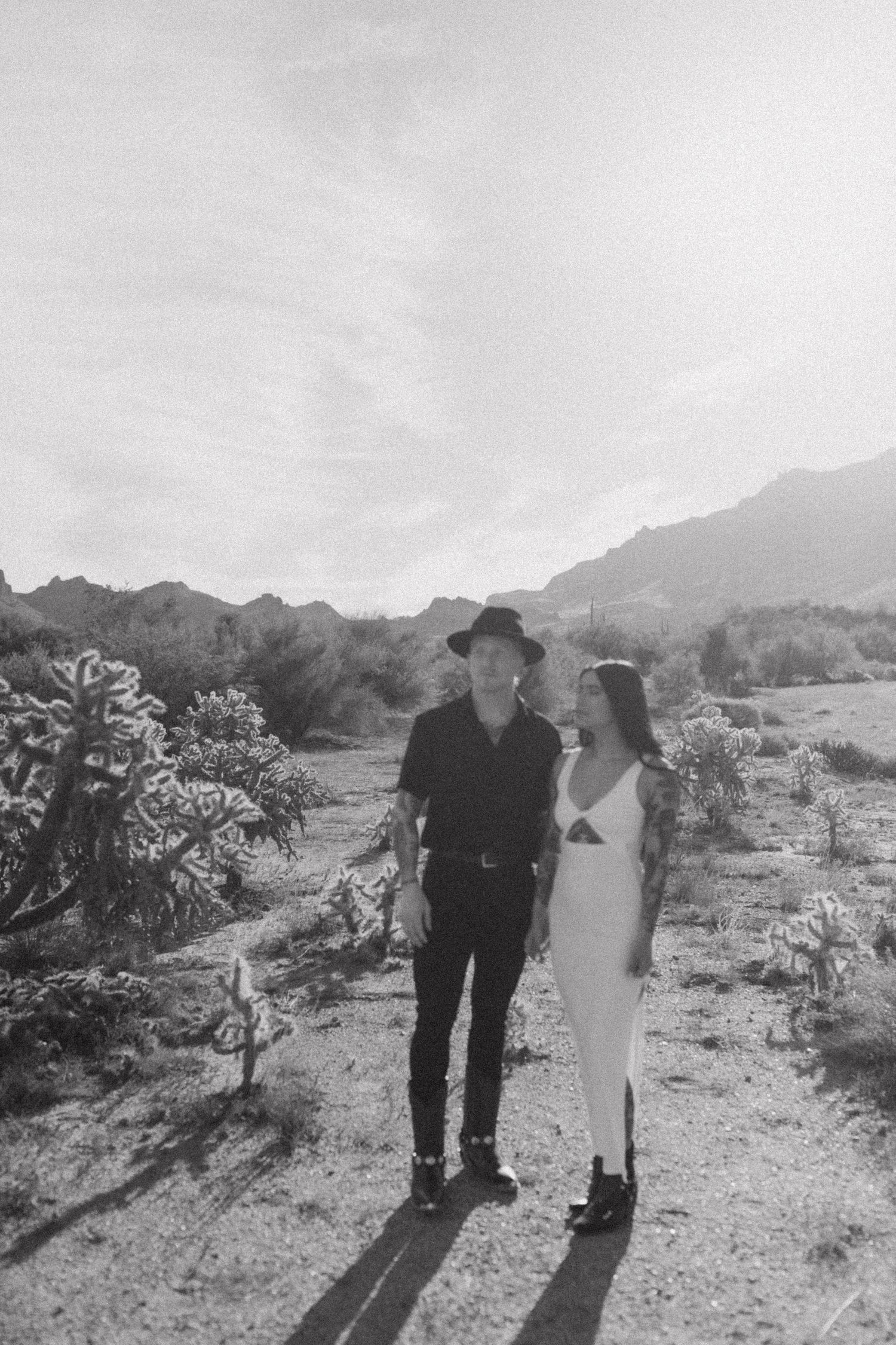 Black and white desert landscape photo shows two figures walking together on a dirt path among desert plants and mountains.