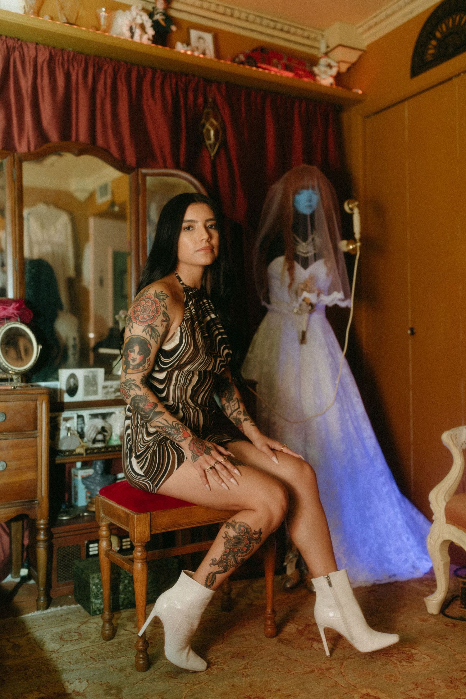 Tattooed person in printed dress and white boots poses in vintage-styled room with wedding dress display in background.