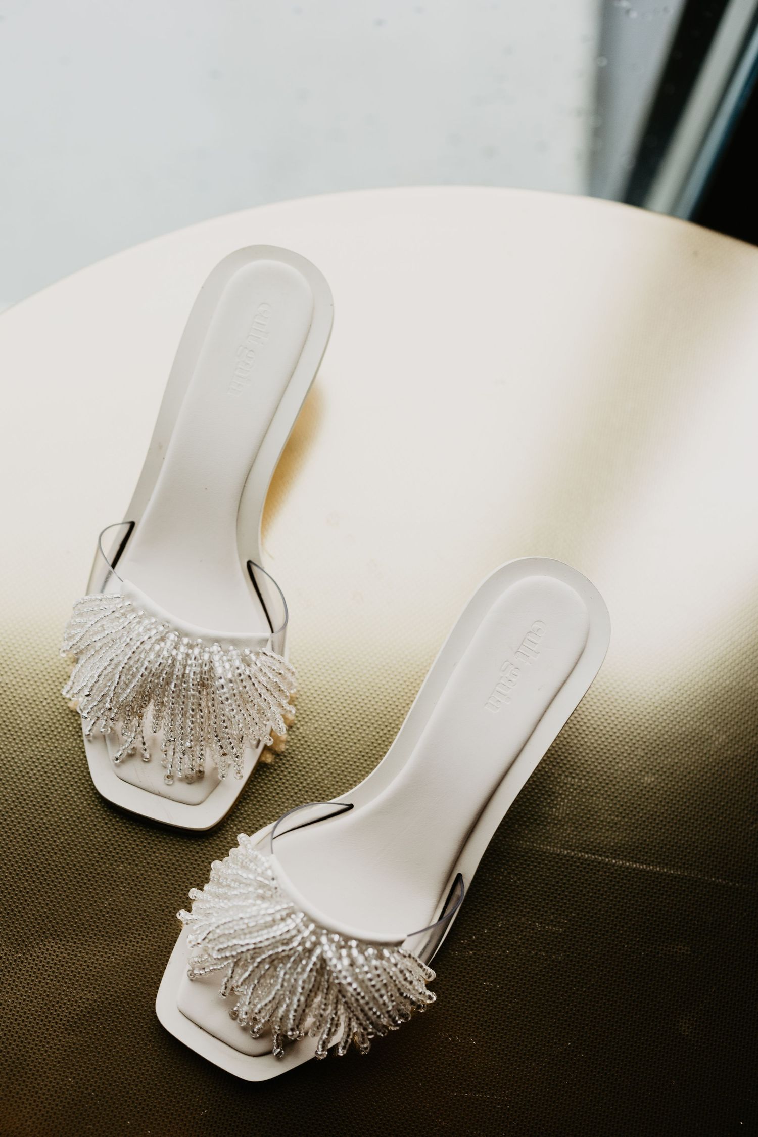 A pair of elegant white bridal slide sandals with delicate beaded fringe details resting on a green surface.