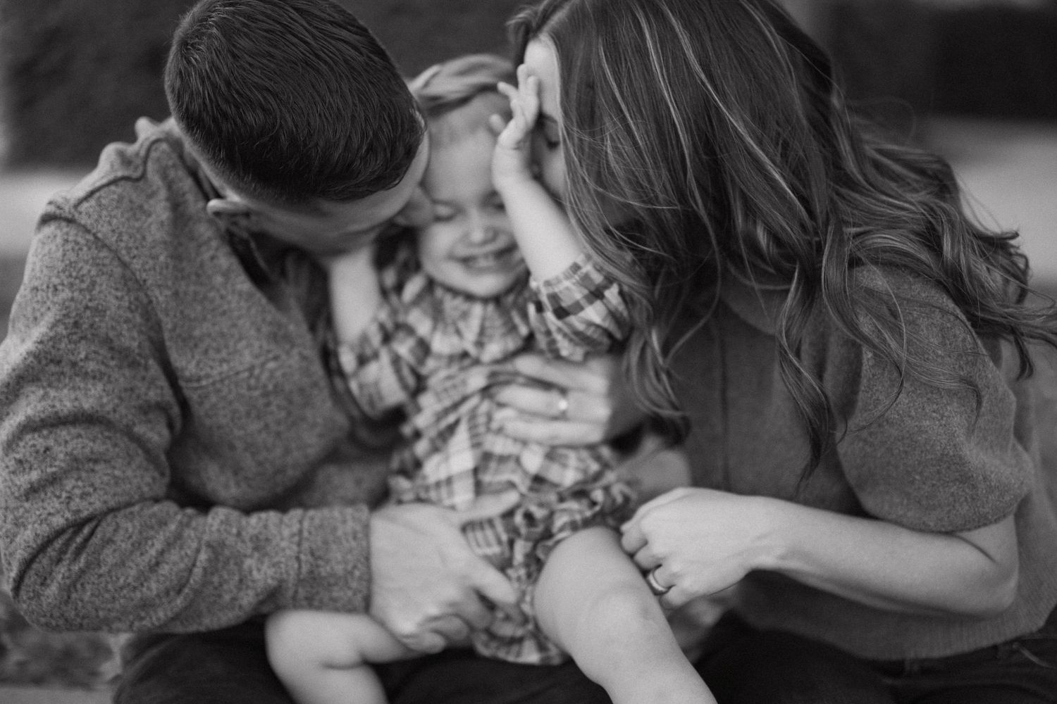 Black and white photo of parents kissing their laughing baby in a tender family moment.