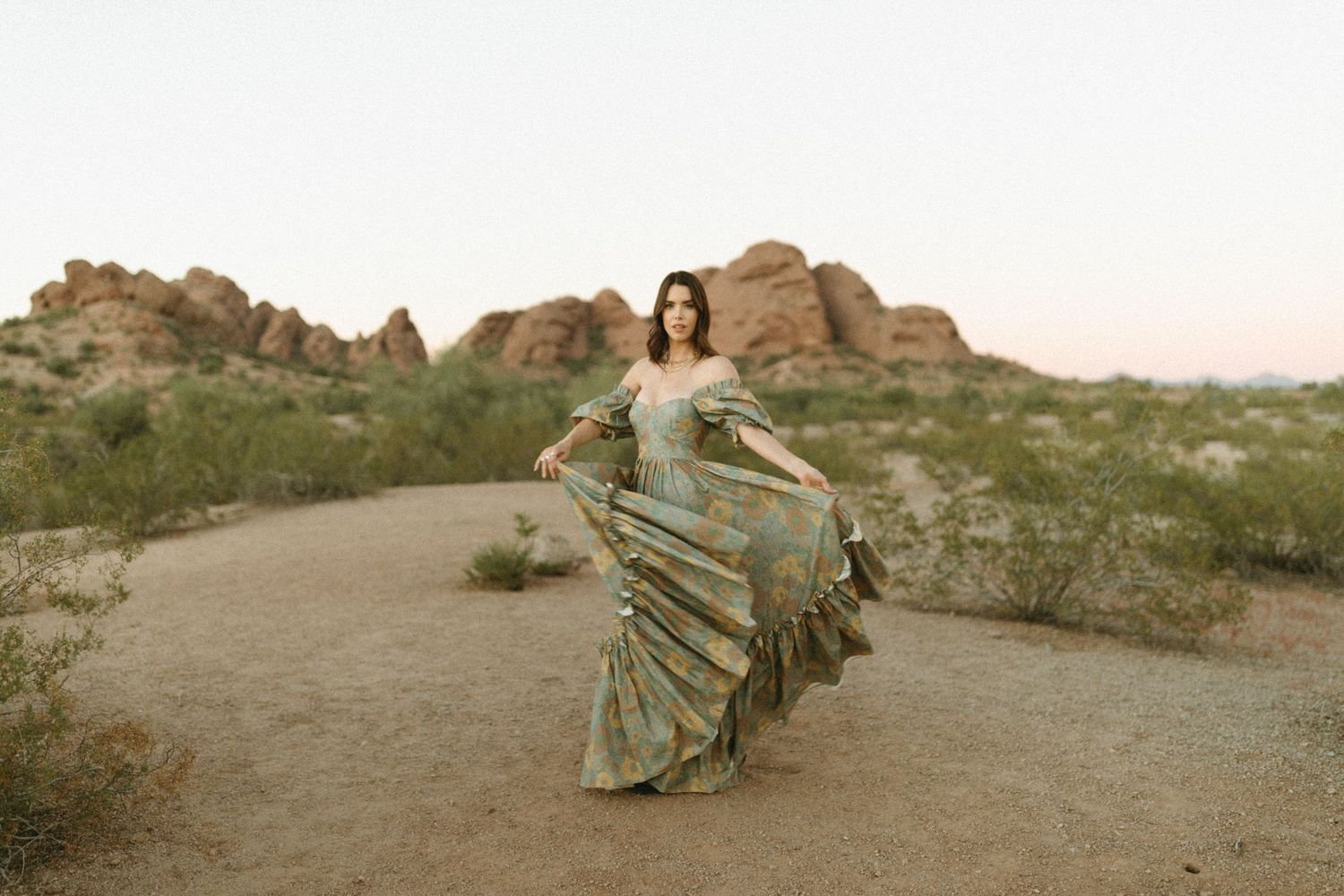 Graceful figure in flowing dress poses against rugged desert landscape during golden sunset hour.