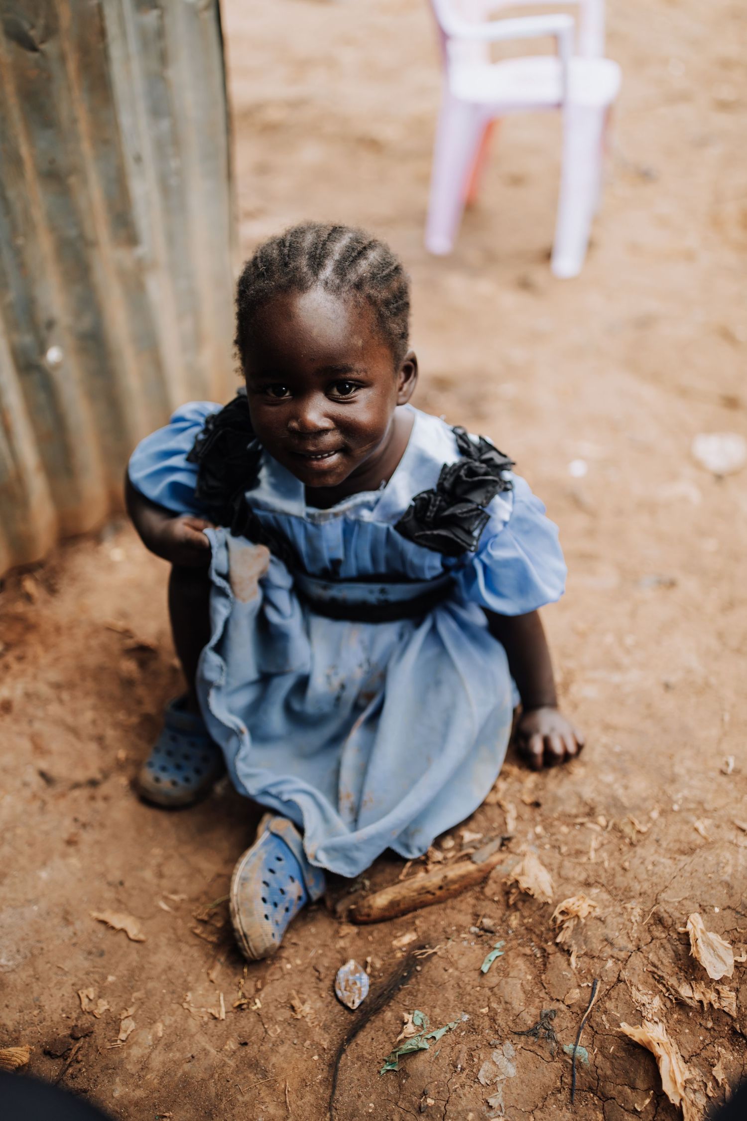 Child wearing a blue dress sits on dirt ground outside with a white plastic chair visible in the background.