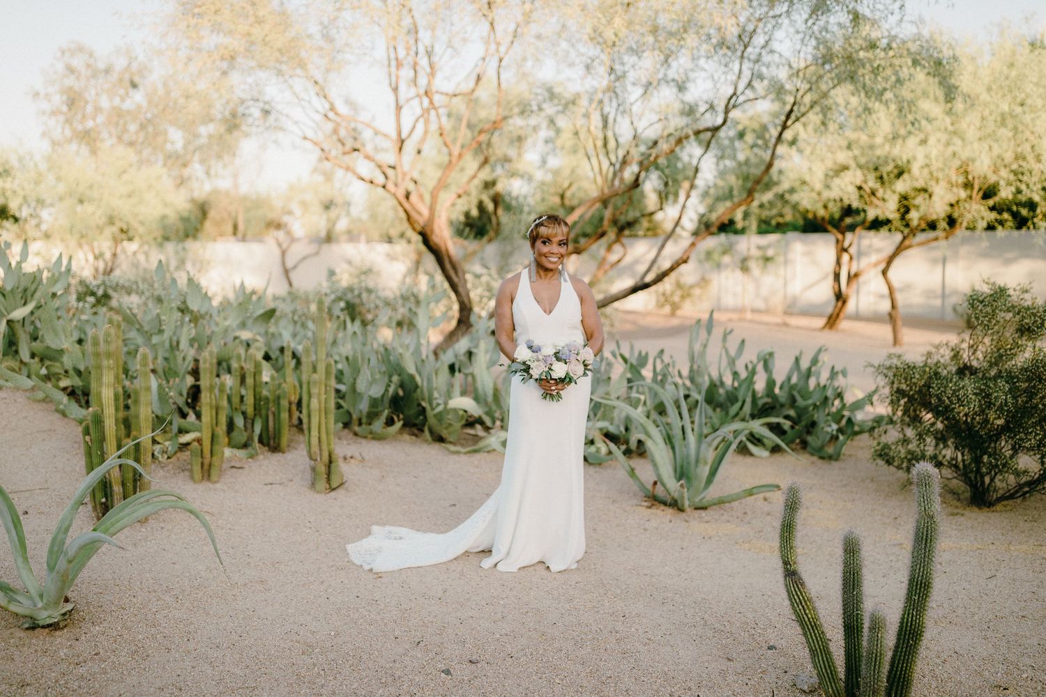 A bride in a flowing white gown stands among desert succulents and agave plants in a natural desert garden setting.