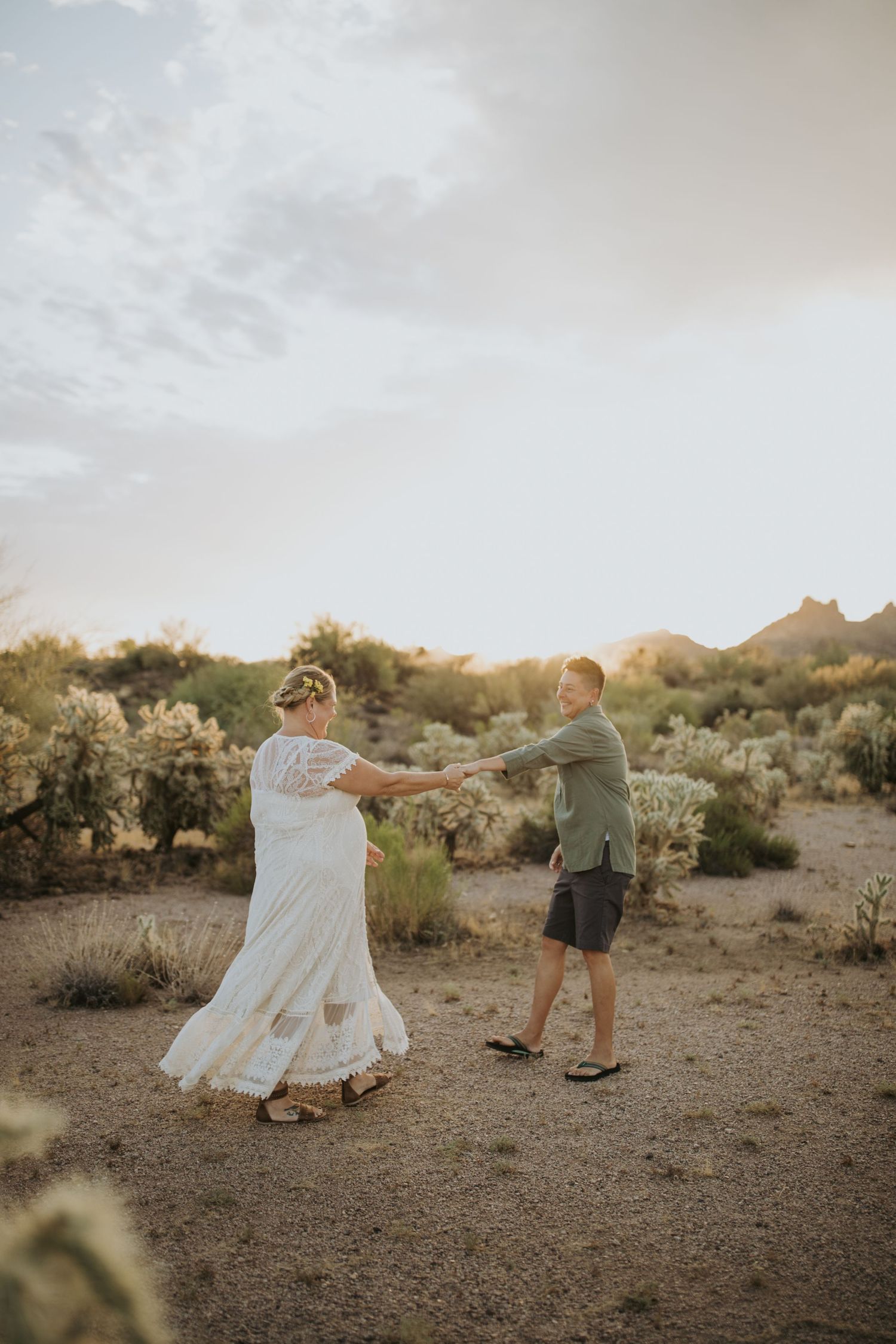 In desert landscape, couple in white dress and casual attire playfully dances and twirls among desert shrubs at sunset.