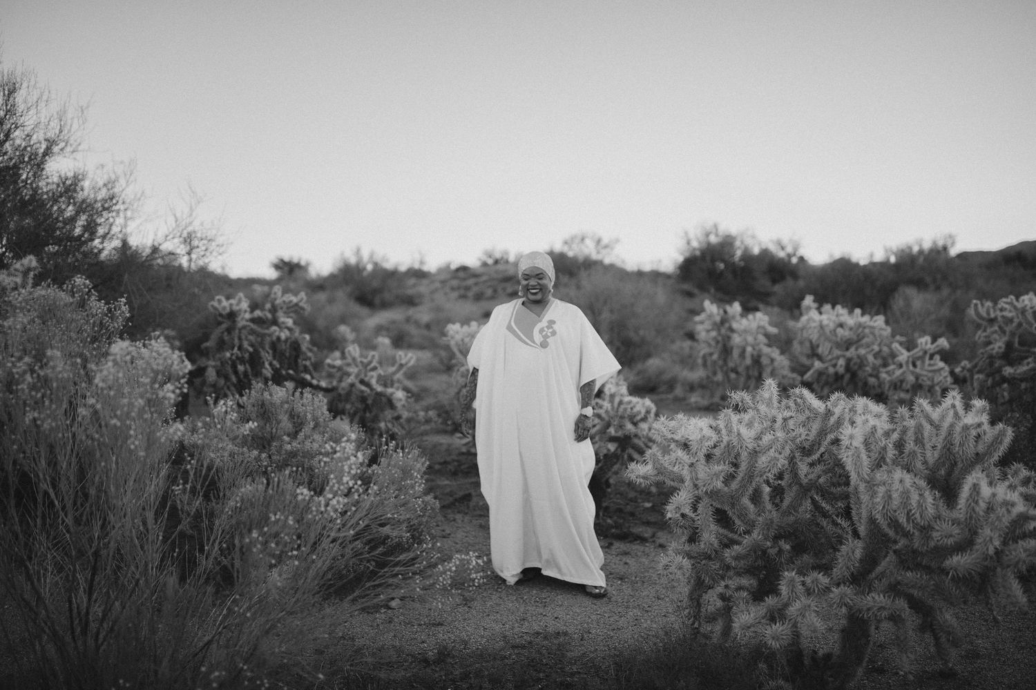 A person wearing a long white robe stands in a desert landscape surrounded by cacti and desert vegetation.