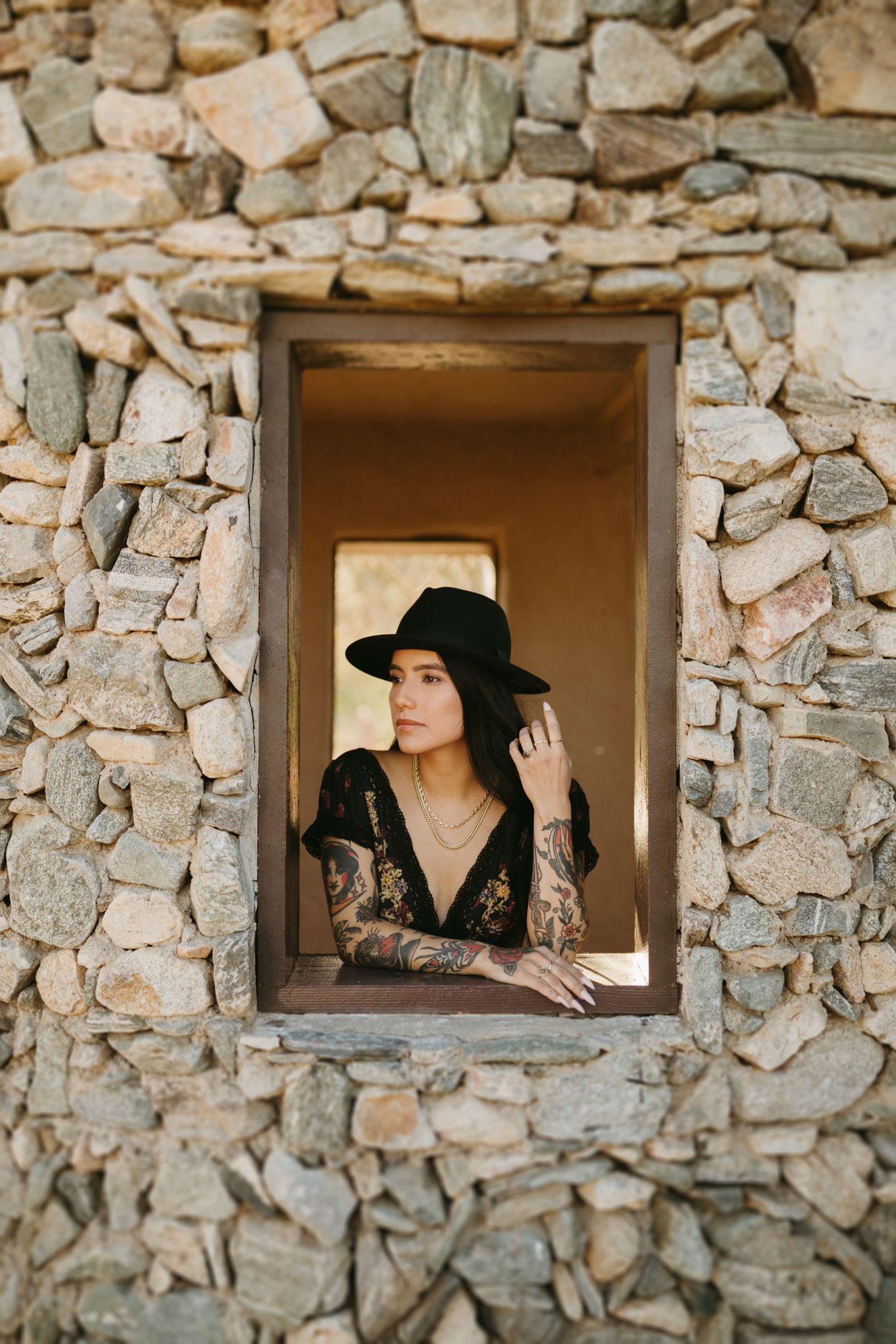 A stone wall window frames a view of someone wearing a black hat and dress with tattoos at sunset.