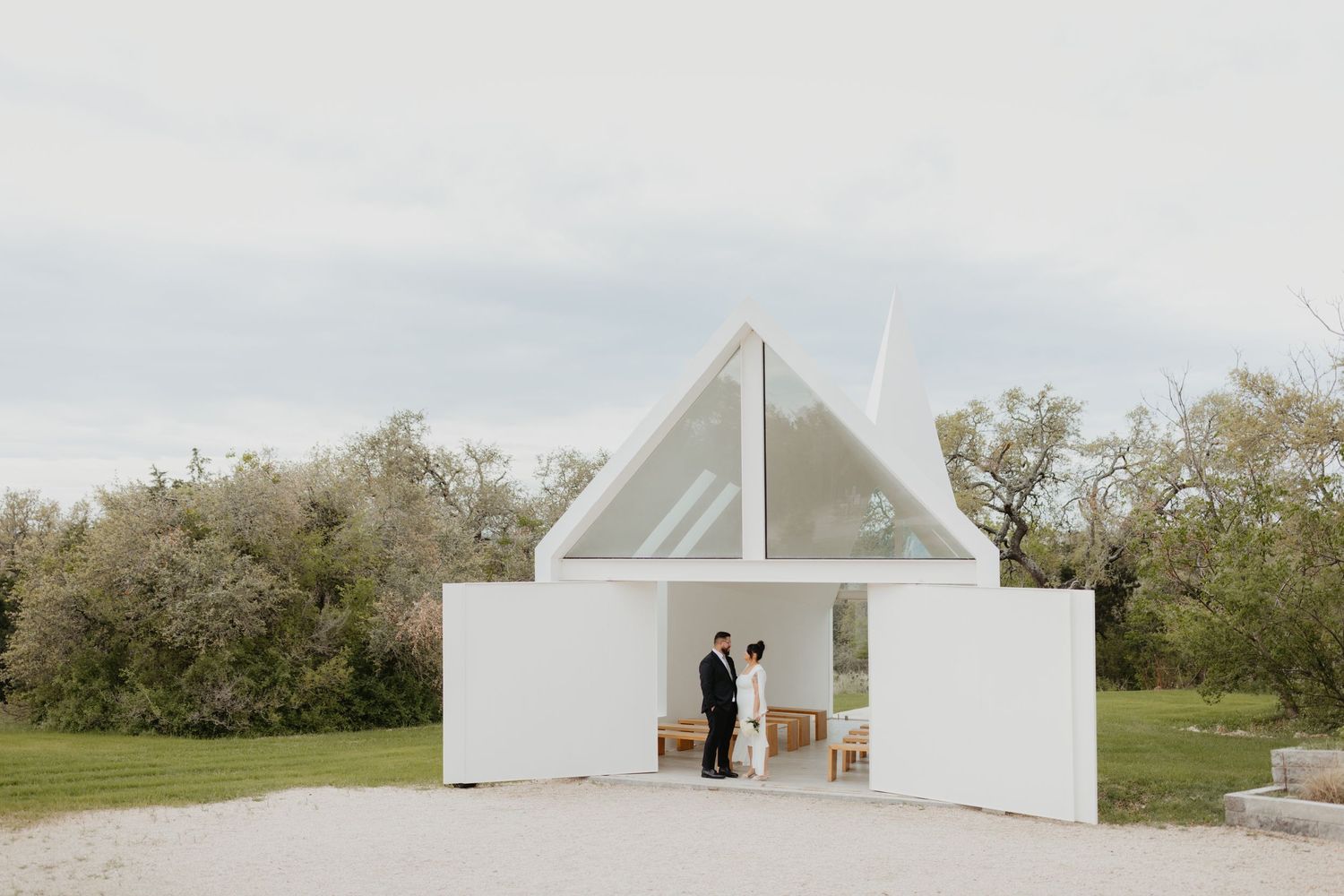 Modern white minimalist wedding chapel stands in a serene garden setting with triangular glass window detail.